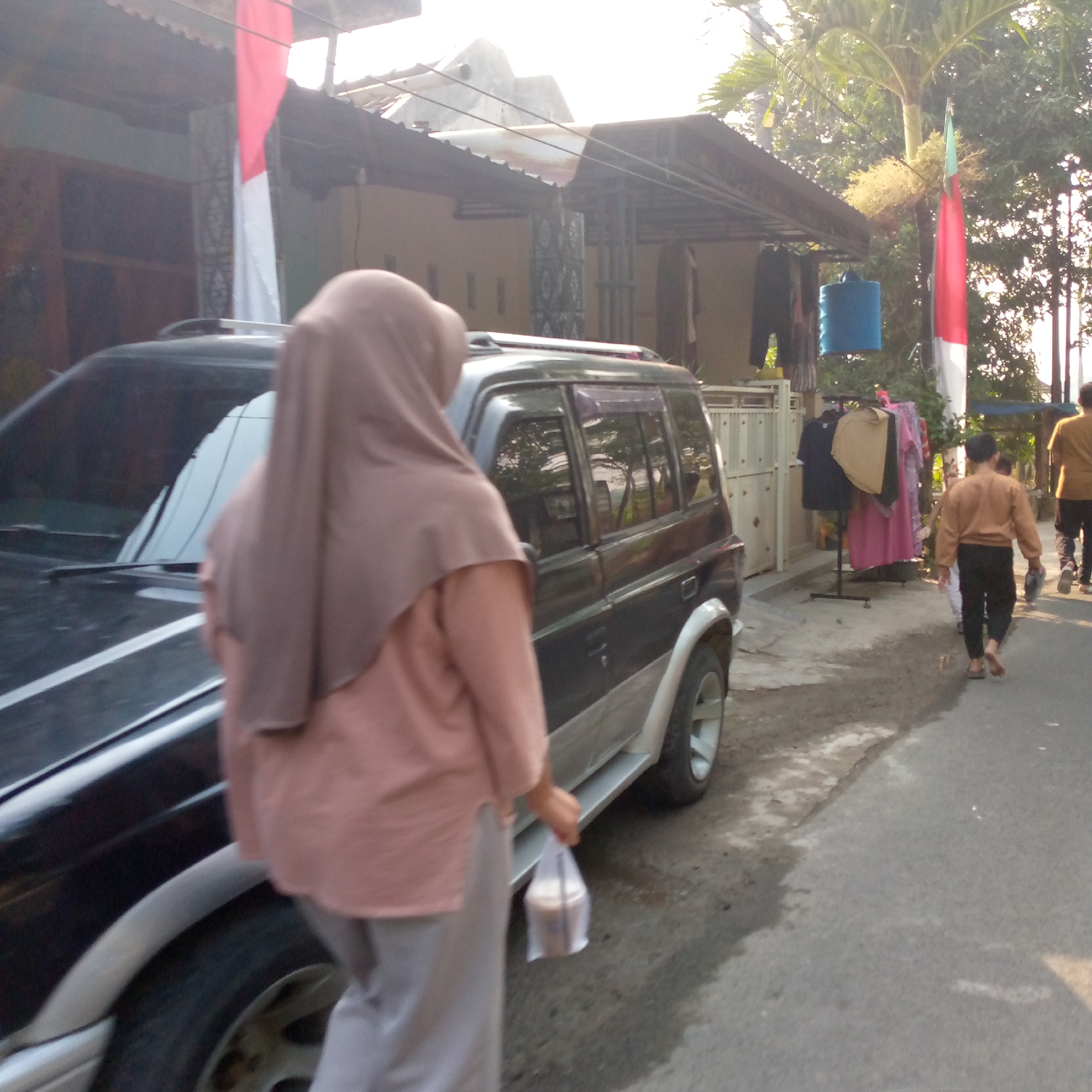 The image depicts a street scene in Tambakagung, Indonesia. In the foreground, a dark-colored SUV is parked, with its rear side visible on the left. A person wearing a light brown hijab and a peach-colored tunic is walking away from the camera, holding a plastic bag containing a beverage in their right hand. They are wearing light gray pants. Behind the SUV, a street is visible with a sidewalk on the left side. Further down the street, a rack with several garments is set up, including a black shirt, a tan top, and a pink dress. A blue water tank is suspended from a pole above the garments. To the right of the garments, an Indonesian flag, red and white, is mounted on a pole. Two children are walking away from the camera towards the right side of the image, one wearing a brown long-sleeved shirt and black pants, and another child is partially visible. In the far right background, another person is walking away, wearing a yellow-brown shirt and dark pants. The environment includes buildings with tiled roofs and vegetation, including a palm tree. The lighting suggests it is daytime, possibly late afternoon, with some shadows present.