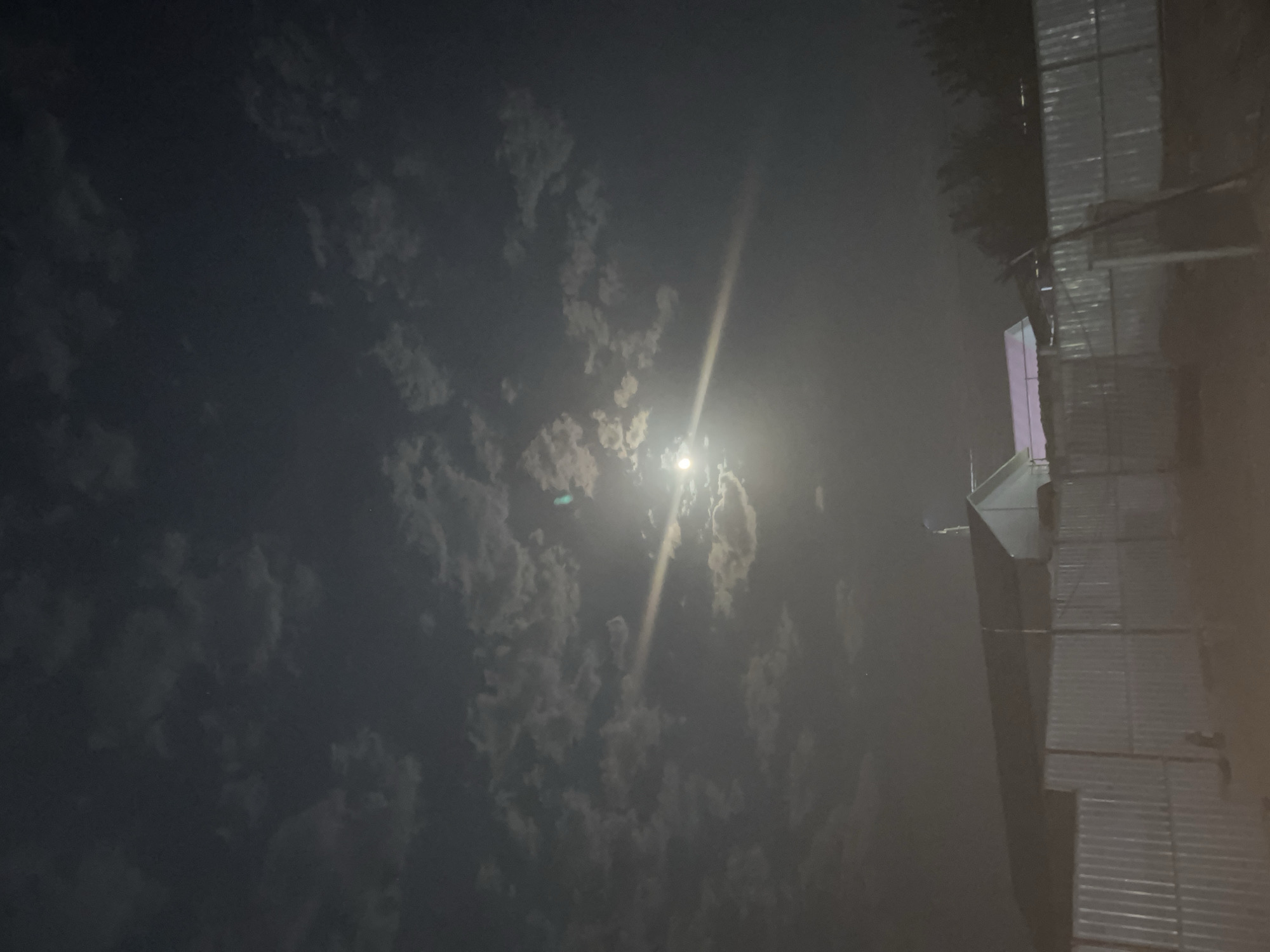 The image captures a nighttime scene in Zarafshan, Uzbekistan, dominated by a bright full moon peeking through scattered clouds. A beam of moonlight cuts across the sky, illuminating the clouds and creating a halo effect around the moon. Below the moonlit sky, a low-rise building with a light-colored, angular roof is visible, partially obscured by a corrugated metal fence that stretches across the foreground. The fence appears to enclose a yard or an industrial area. Trees and other structures are dimly silhouetted in the background. The overall atmosphere is serene and nocturnal, with the artificial light from the building offering a subtle contrast to the natural luminescence of the moon.