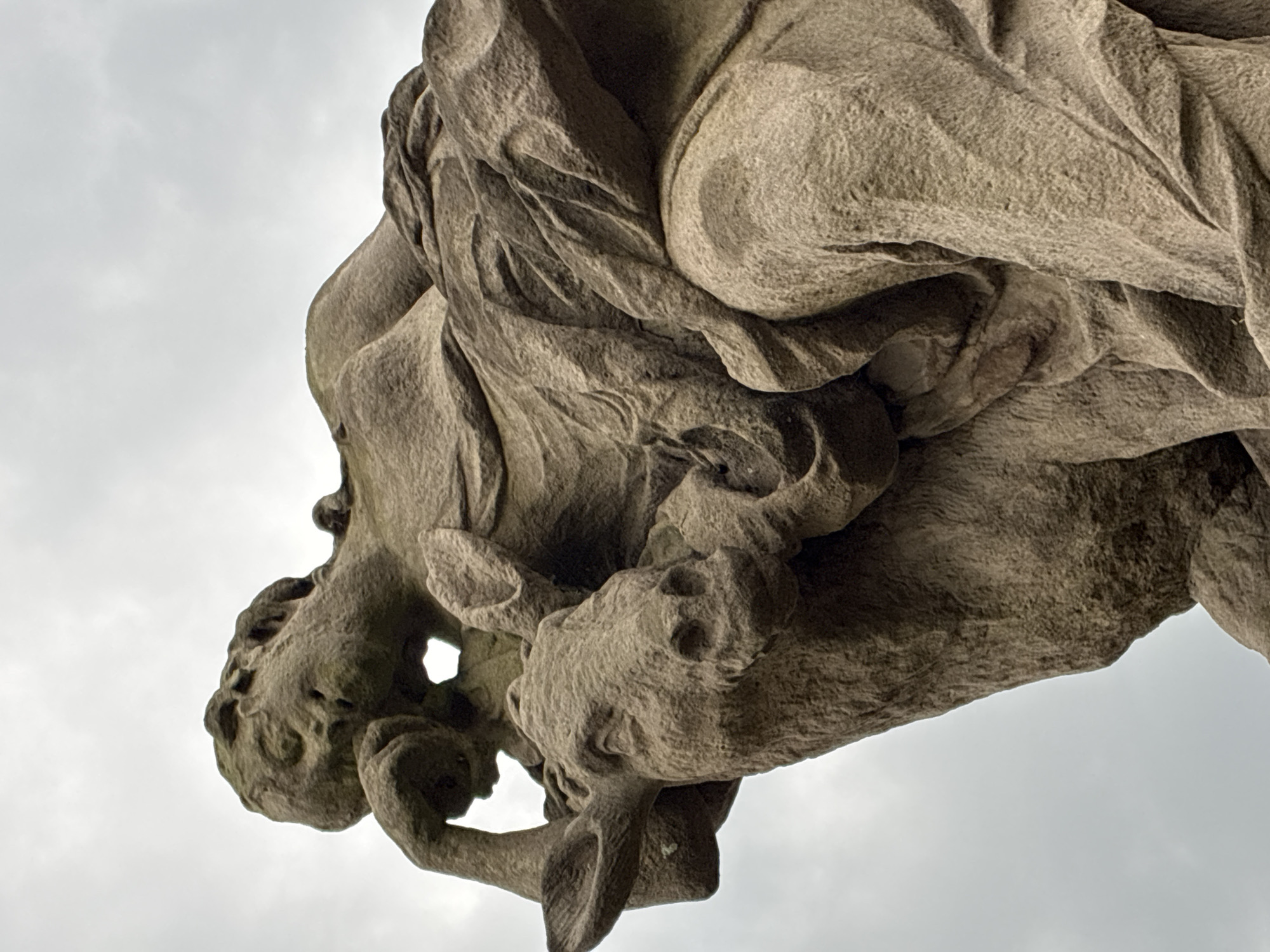This is a close-up, low-angle photograph of a weathered stone sculpture in Kuks, Czechia, depicting a figure with a deer. The sculpture is made of rough-hewn stone, exhibiting signs of age and erosion. The figure appears to be leaning back, with its right arm resting on its head and its face turned upwards. The figure is draped in flowing garments that are intricately carved. A deer's head is visible in the foreground, positioned to the left of the figure's head. The deer's ears are perked, and its eyes are open. The background consists of an overcast, pale sky. The lighting is diffused, creating soft shadows that highlight the textures and contours of the stone. The image focuses on the upper portion of the sculpture, emphasizing the detailed carving and the weathered patina of the stone.