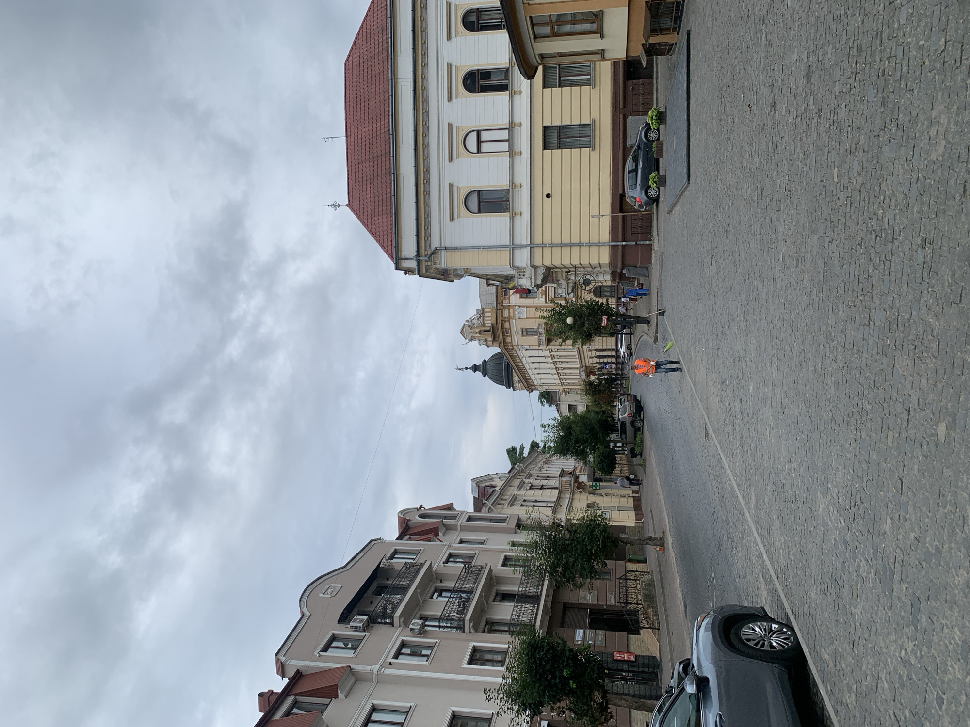 The image depicts a cobblestone street in Kolomyia, Ukraine, on a cloudy day. The street is lined with buildings, some of which are ornate and feature red tile roofs. A man in an orange vest and blue pants is sweeping the street, holding a broom. Several cars are parked along the side of the street. The overall atmosphere appears to be quiet and ordinary, possibly early morning or late afternoon.