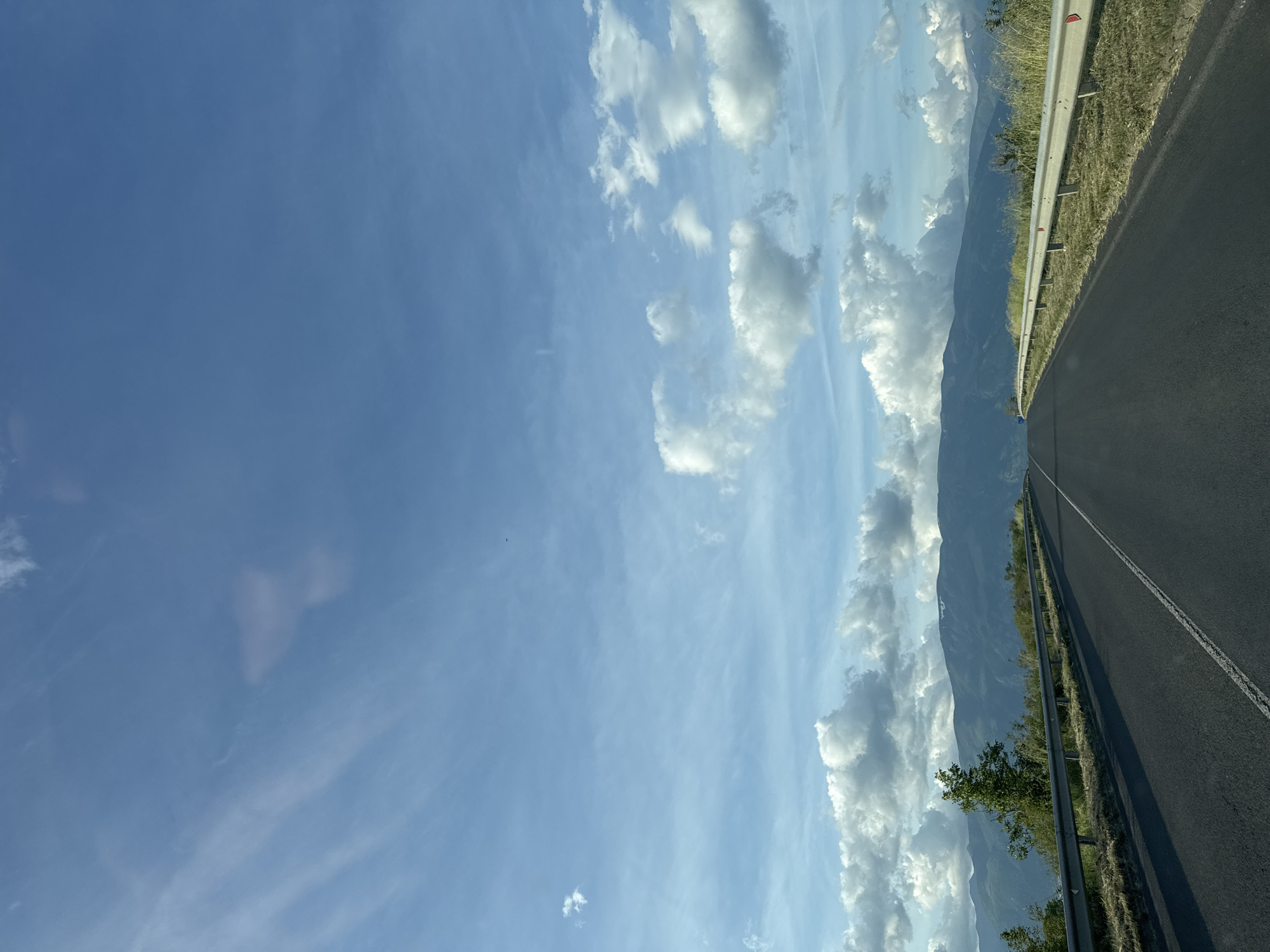 A vertically oriented photograph shows a highway with a mountainous backdrop and a partly cloudy sky. The highway, made of black asphalt, has a white dashed line dividing it into two lanes. A silver metal guardrail runs alongside the road's edge, and beyond the guardrail, there is grassy vegetation. In the distance, a mountain range is visible. The sky is predominantly blue with scattered white clouds.