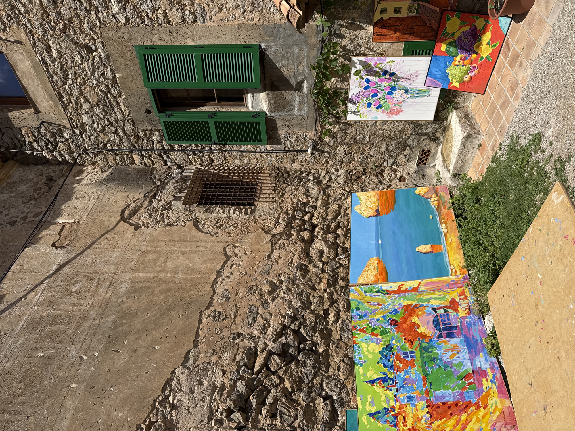 The image shows an alley in Valldemossa, Spain. The alleyway walls are constructed of a mix of bare stone and plaster surfaces. A window with green shutters is visible on the left side. A metal grate is located beneath the window. Several paintings are displayed against the wall on the right side of the alley, as well as on the ground. The ground is composed of tiled and stone surfaces with patches of green vegetation. There are no people visible in the image.