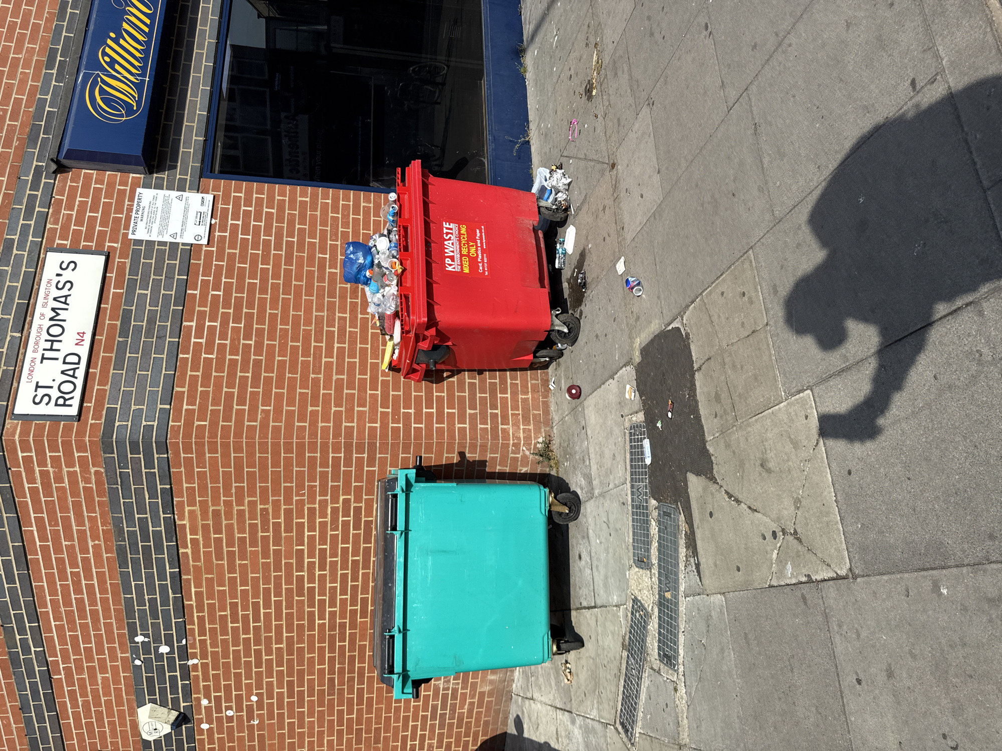 The image shows a street scene in the London Borough of Islington, United Kingdom, specifically on St. Thomas's Road (N4 postal code). A red brick building occupies the left side of the frame, featuring a "William" sign and a "Private Property" notice. Two waste bins, one red and overflowing with recyclable materials, the other teal, are positioned on the sidewalk. Litter is scattered around the red bin. The sidewalk is concrete and has several drain covers. The shadow of a person is visible on the sidewalk.