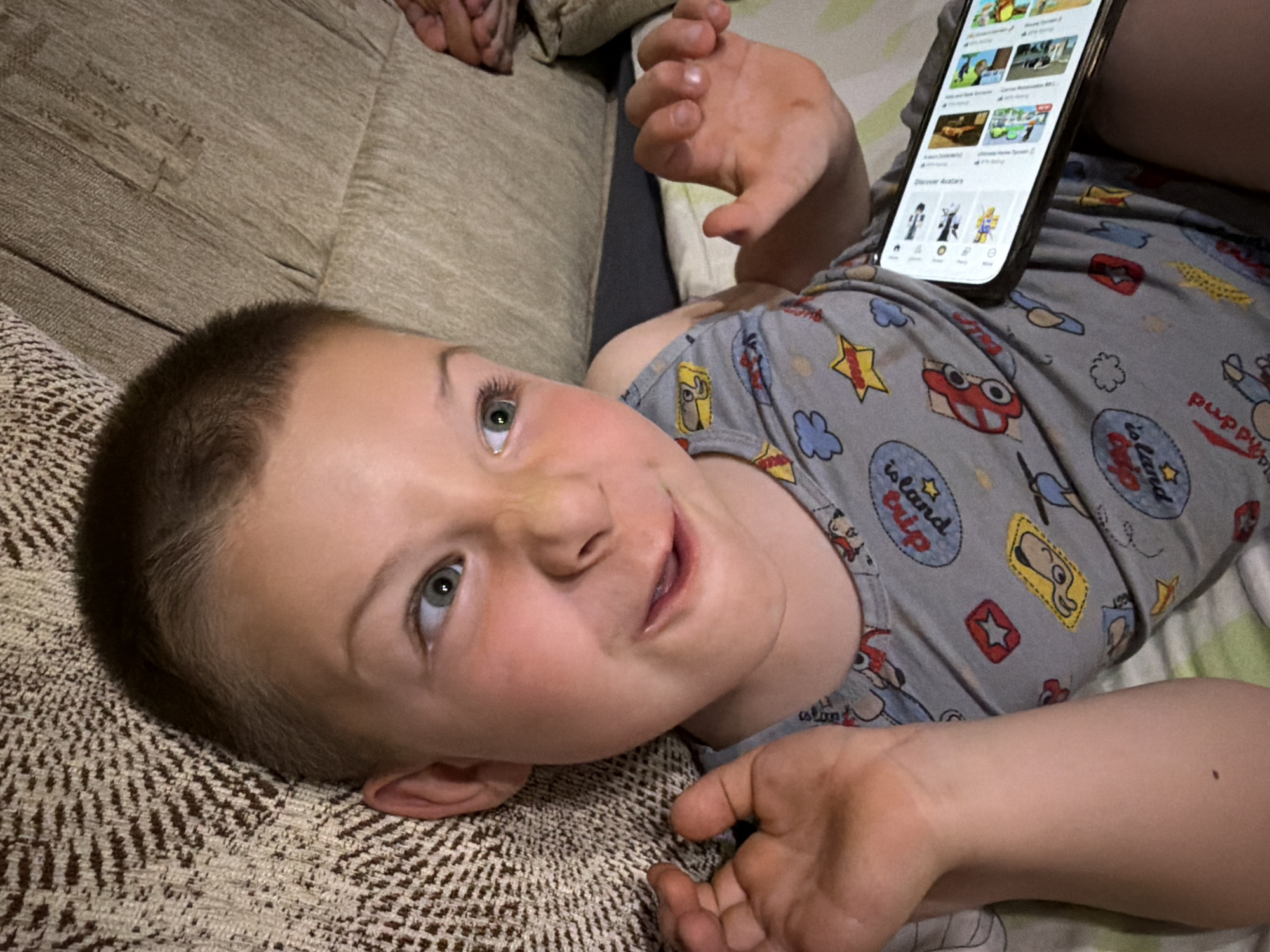 A young boy with short brown hair and green eyes is lying down and looking to his right with a playful expression. He is wearing a grey sleeveless shirt with a pattern of cartoon cars, clouds, stars, and the text "Fun" and "learn" on it. His arms are bent at the elbows, with his left hand raised near his face and his right hand outstretched. A smartphone is resting on his shirt, displaying a grid of images and text, indicating he is likely watching something or browsing on the device. The background consists of a textured, brown couch. The scene appears to be indoors, possibly in a living room. The lighting suggests it is evening or nighttime. The provided context places this scene in Zmeitsa, Bulgaria, though no specific landmarks or city details are visible in the image itself.