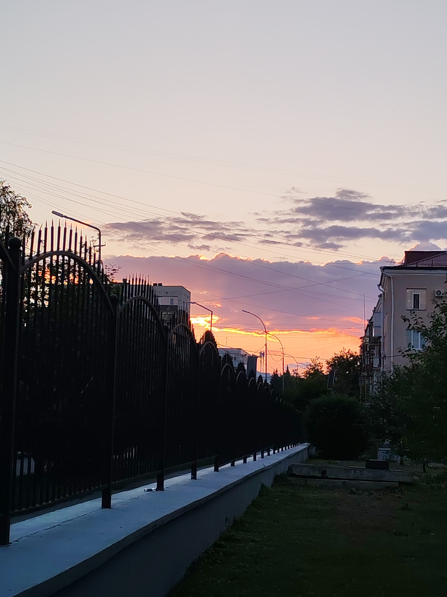 A sunset sky with orange and purple hues is visible above a city skyline. A black metal fence runs across the foreground, with a white wall beneath it. Buildings and trees are visible behind the fence. Overhead, power lines crisscross the sky. The scene is set in an urban environment.