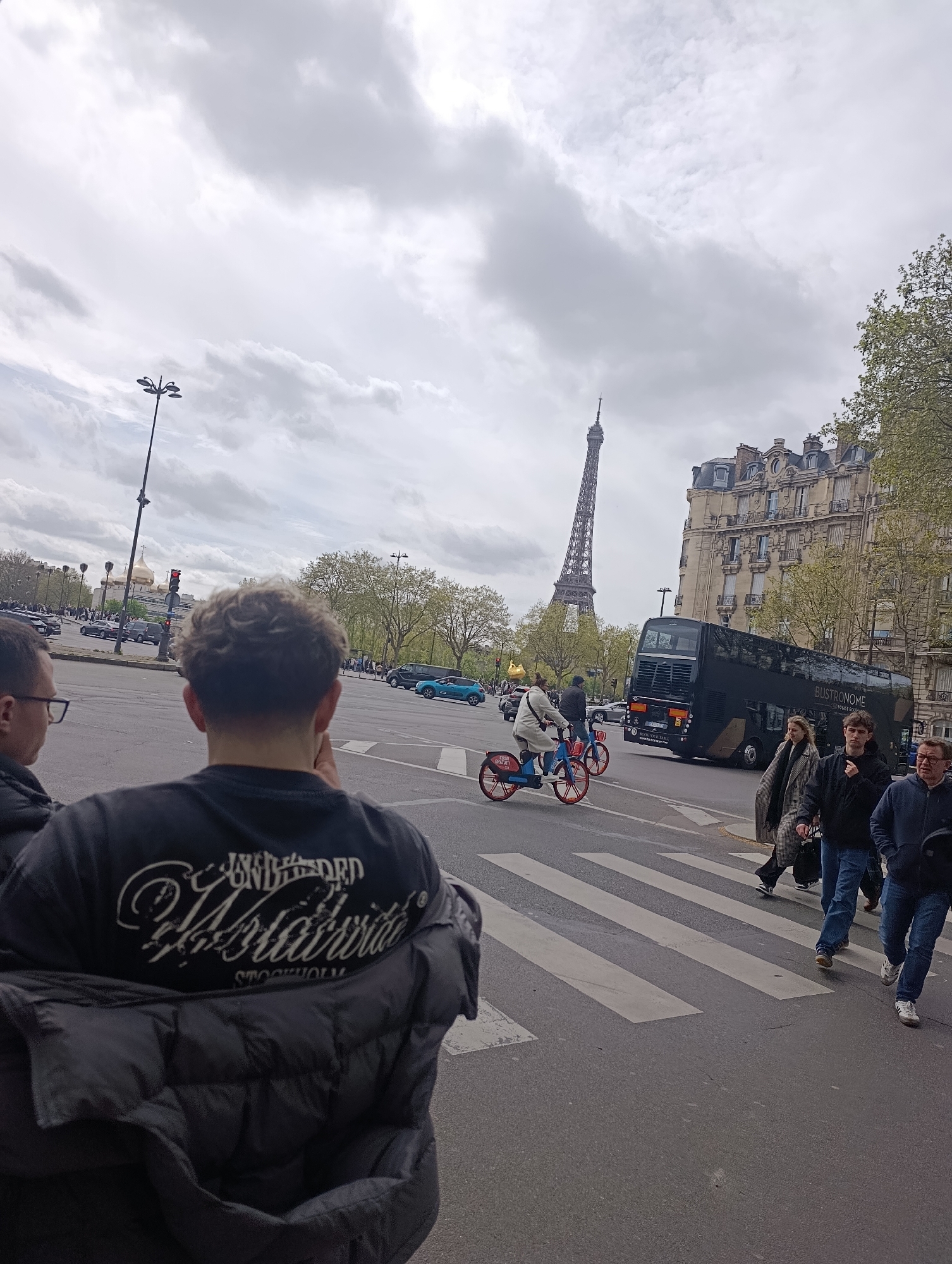 The image captures a street scene in Paris, France, with the Eiffel Tower visible in the background. In the foreground, a person wearing a black t-shirt and jacket stands with their back to the camera. To their left, another person is partially visible. Several individuals are crossing the street on a pedestrian crossing. Two people are riding bicycles. A black double-decker bus, identified as "Bustronome," is present on the street. Buildings line the street, and trees are visible. The sky is overcast. Traffic lights are visible in the background.
