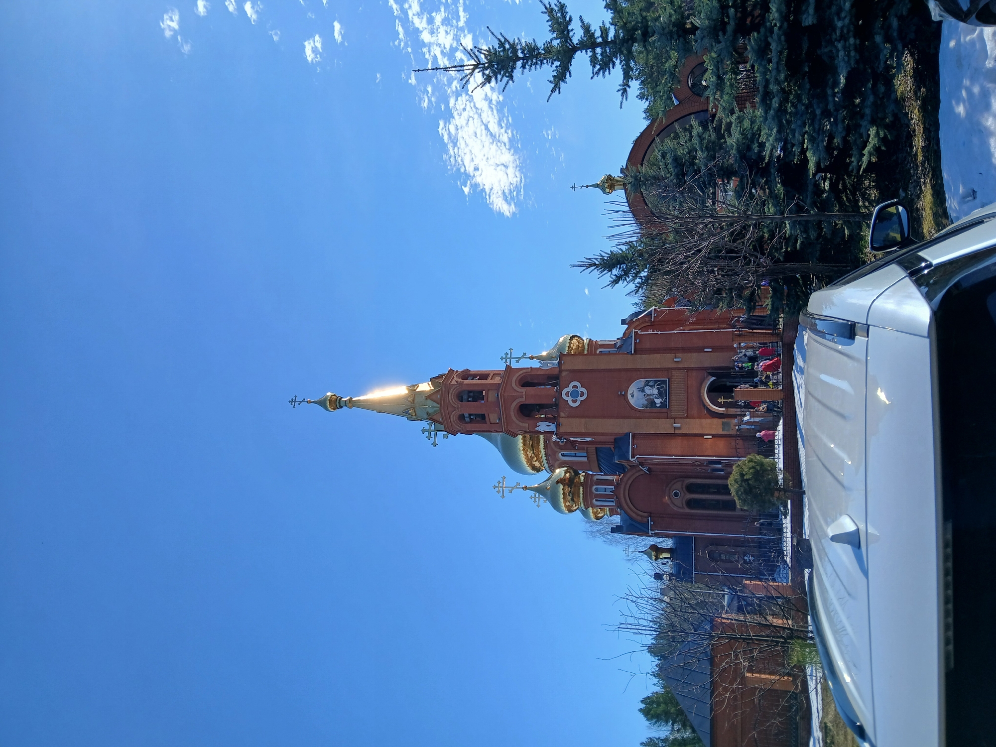 The image shows the Church of the Nativity of Christ in Chaikovsky, Perm Krai, Russia. The church is made of red brick and has several golden domes and crosses. The sky is blue with some clouds. Part of a white car is visible in the foreground. There are trees surrounding the church. People are near the church.