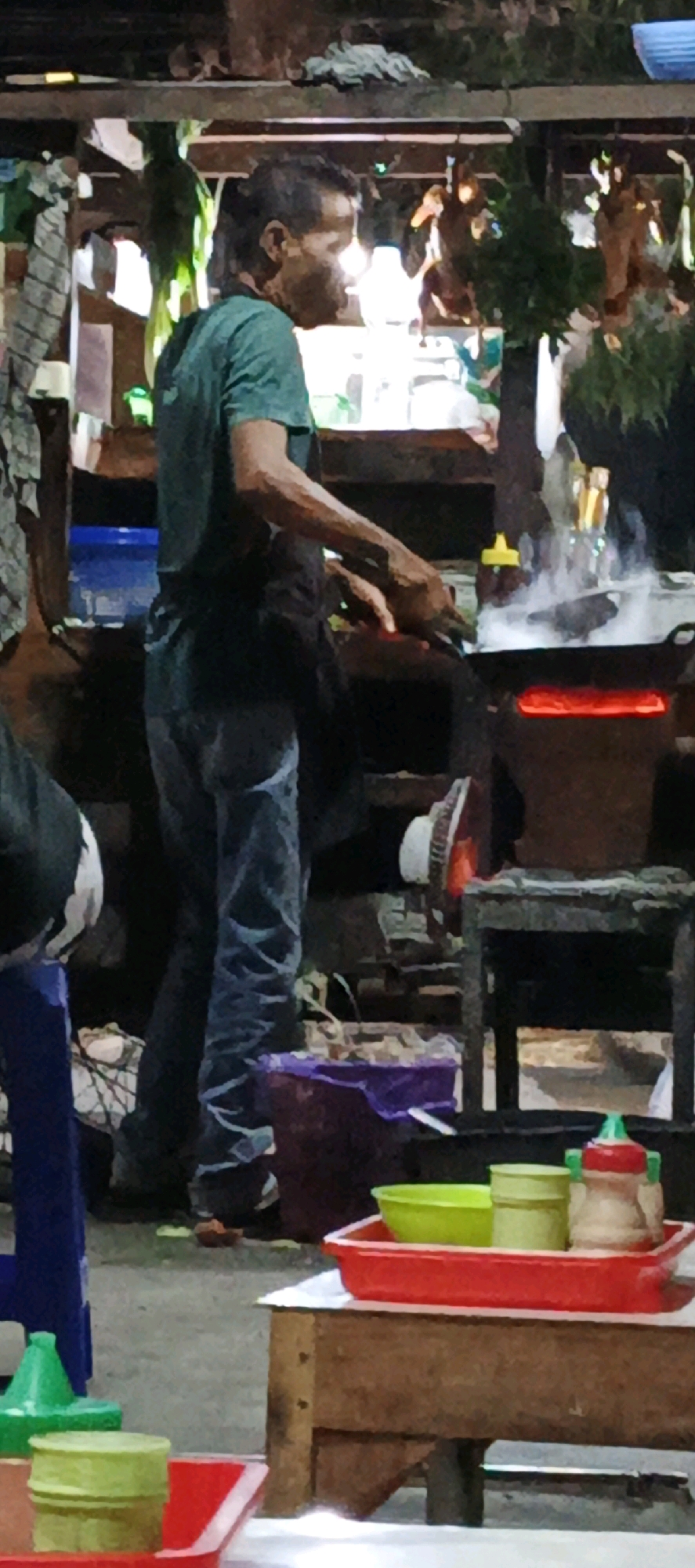 The image depicts a man in Sidoagung, Indonesia, cooking at an outdoor food stall. He is wearing a green t-shirt and blue jeans, and appears to be stirring food in a wok over a fire. The stall is set up with various cooking utensils and ingredients, including bottles and containers of sauces and condiments. In the foreground, a red tray holds a green bowl and a yellow container, possibly for serving food. The overall scene suggests a bustling street food environment.