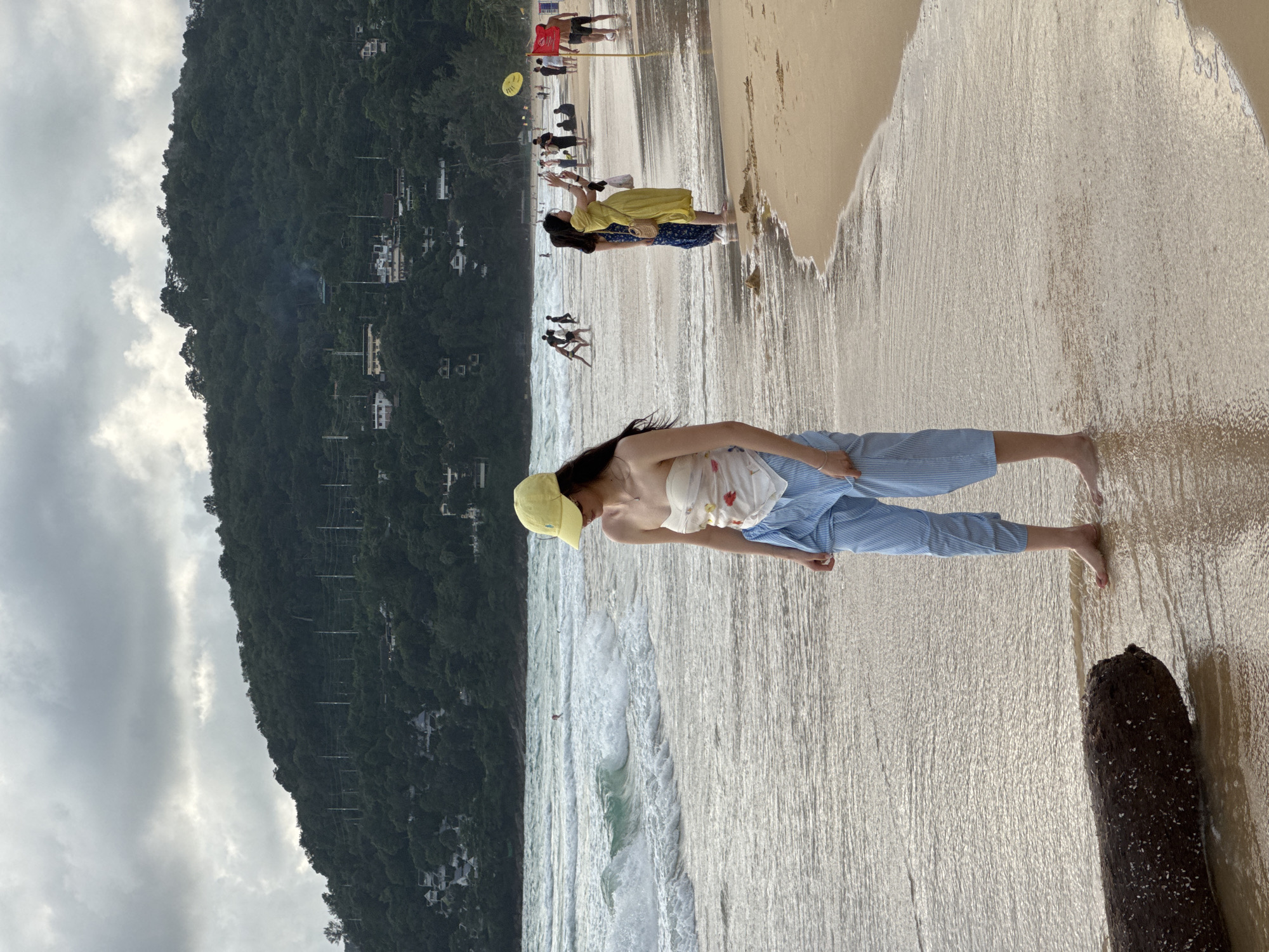 A young woman wearing a yellow baseball cap, a strapless white top with floral accents, and blue-and-white striped pants stands at the edge of the ocean on a sandy beach. She is looking down and to her right. Her feet are submerged in the shallow water. A large, dark log lies on the sand to her left. The background shows choppy, light-colored waves rolling onto the shore, and further back, a green, tree-covered hillside with scattered buildings and power lines. Several other people are visible in the distance, some wading in the water and others walking on the beach. A yellow flag and a red flag are planted in the sand further down the beach. The sky is overcast with a mix of gray and white clouds. This scene is located in Karon, Thailand.