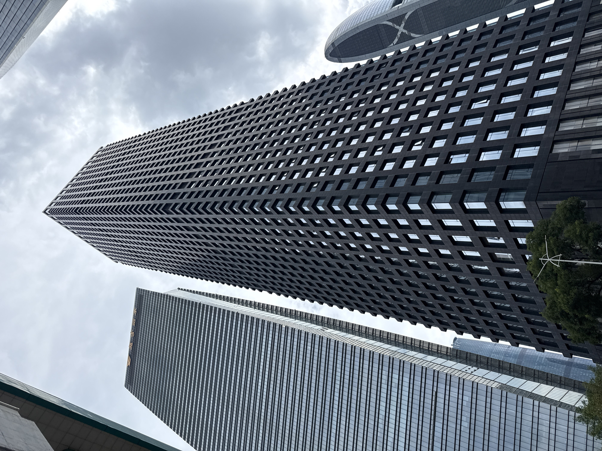 An extreme low-angle, full shot captures a cluster of towering skyscrapers in Tianhe District, China, under a cloudy sky. The most prominent structure is a dark, rectangular skyscraper occupying the center of the frame. Its facade is a grid of dark panels and small, reflective windows, creating a textured appearance. To the left, a taller, slender skyscraper with a glass facade is visible, its surface reflecting the sky and adjacent buildings. Its upper section bears golden Chinese characters. To the right and slightly behind, a uniquely shaped, curved skyscraper with a patterned facade adds a distinct architectural element to the skyline. The bases of the buildings are partially obscured by lush green trees and a white street lamp with a circular element. The composition emphasizes the immense scale and modern architecture of the urban environment.