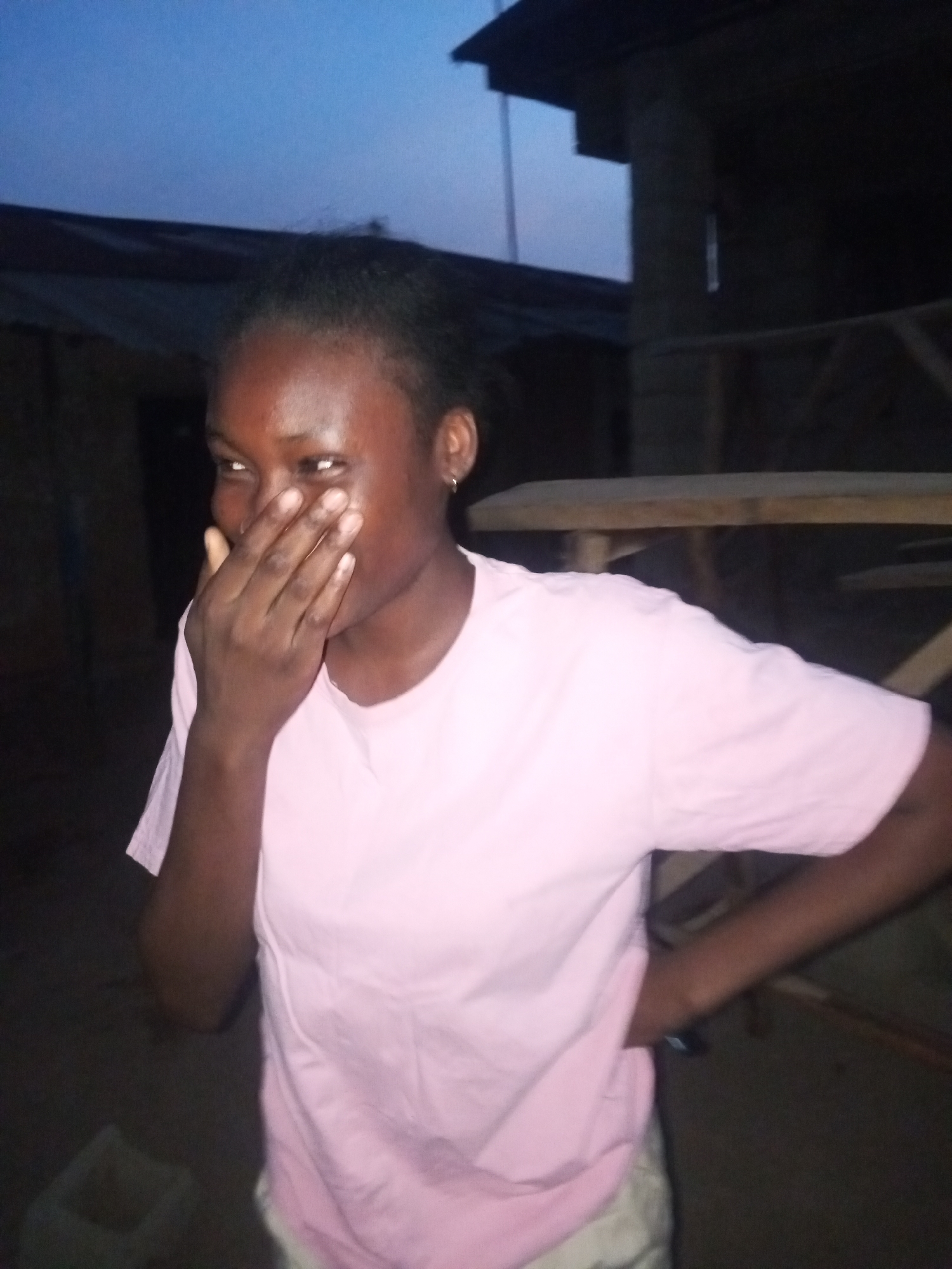 A young woman with dark skin wears a light pink t-shirt and light-colored shorts. She has an earring in her left ear and is covering her mouth with her right hand. She is smiling with her eyes. Her left hand is on her hip. The photo appears to have been taken outdoors at dusk or dawn, with a structure visible behind her that seems to be a building under construction or a simple dwelling. The location appears to be a rural or semi-rural area. The location is stated as Bukuru, Nigeria, but the photo itself does not provide explicit location details.