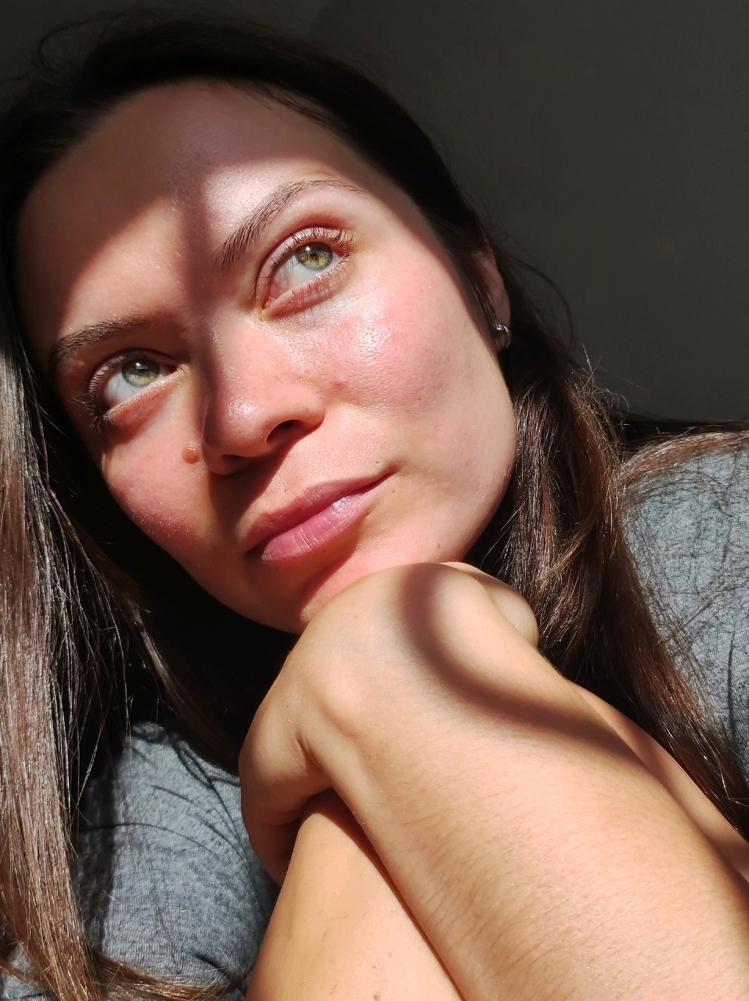 A close-up, low-angle, portrait-style photograph captures a young woman's face as she gazes upward and to her right. Sunlight casts dramatic shadows across her forehead, cheekbone, and nose. Her left eye, a striking green, is fully visible and reflects light. Her lips are slightly parted, and her chin rests on her folded right hand, which is positioned in front of her chest. Her brown hair cascades down her shoulders and partially obscures the grey fabric of her t-shirt. The background is dark, with the exception of some light filtering from the upper left corner, suggesting an indoor setting with a light source to the side. A small, raised mole is visible on her right cheek. The woman is wearing a small hoop earring in her left ear. There is no discernible indication of the city of Chaikovsky, Russia, within the visual content of this image.