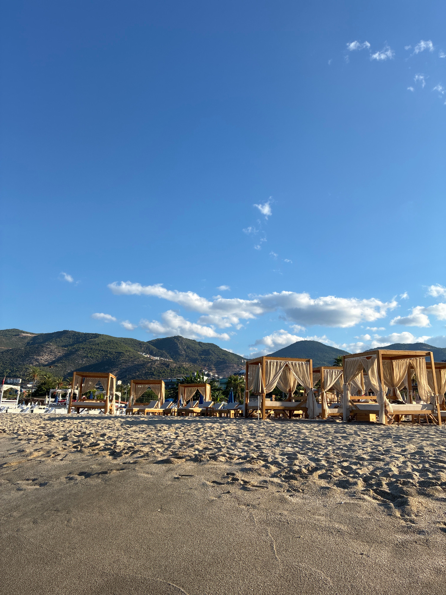 A vertical, eye-level shot captures a serene beach scene on a sunny day in Alanya, Turkey, with a clear blue sky dominating the upper portion of the frame. Scattered, fluffy white clouds drift lazily across the sky, adding a touch of softness to the expanse.
In the middle ground, a row of inviting, wooden cabanas with sheer white curtains are set up on the sandy beach. These cabanas, arranged in a line, appear to offer a private and shaded retreat for beachgoers. Some of the cabanas are occupied by people relaxing on lounge chairs, visible through the curtains. To the left and right of the cabanas, there are more beach chairs and umbrellas, indicating a well-equipped resort or public beach area.
The background is characterized by a majestic mountain range, covered in lush green vegetation. The slopes of the mountains are dotted with buildings, suggesting a town or resort built into the hillside, characteristic of Alanya's landscape. The sunlight casts a warm glow on the sand and the mountains, creating a peaceful and idyllic atmosphere.
The foreground is dominated by the textured sand of the beach, with footprints and undulations suggesting recent activity. The overall scene conveys a sense of relaxation, vacation, and natural beauty, typical of a Mediterranean coastal destination.