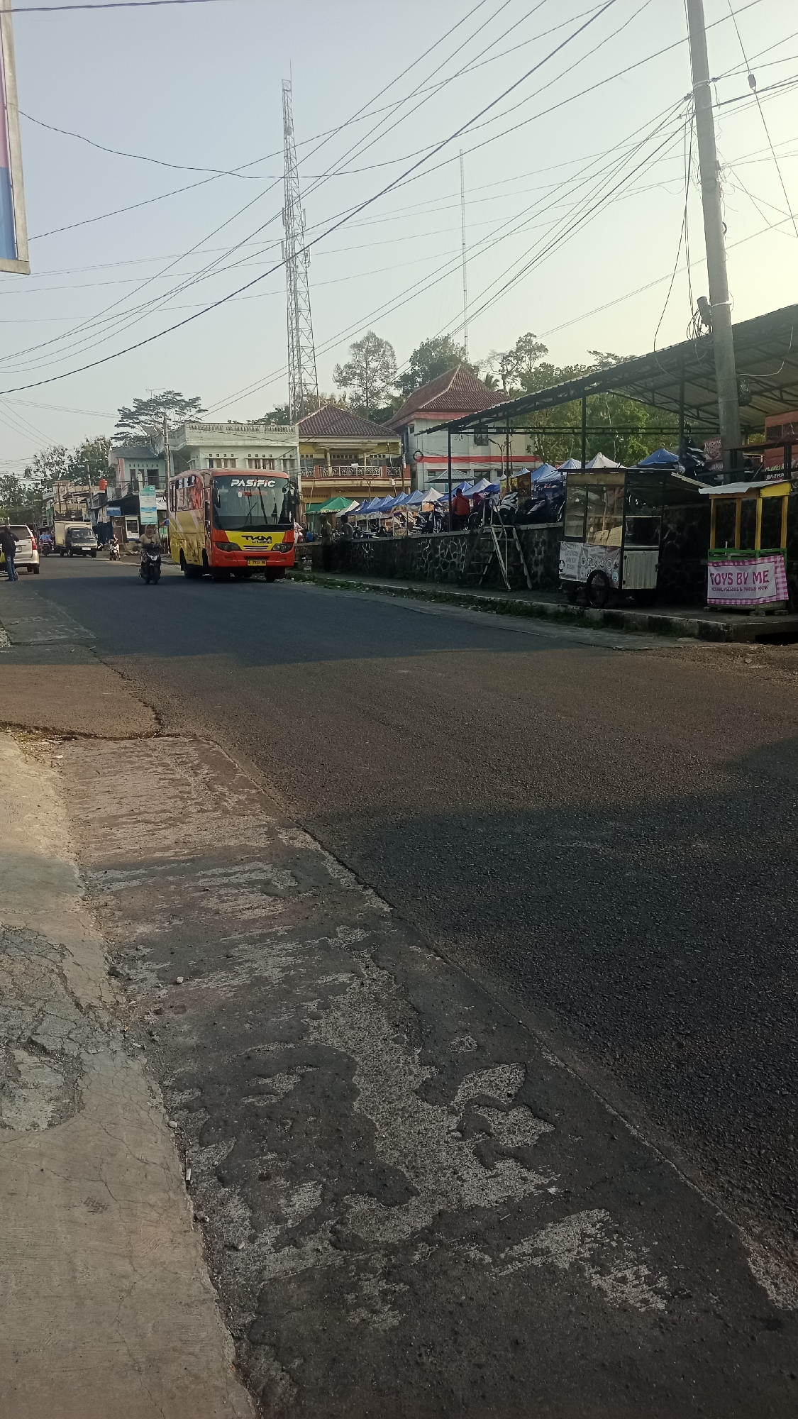 The image depicts a street scene in Pakemitan, Indonesia, likely a commercial area. A yellow and red bus, with "PASIFIC" written on its front, is driving down the road. Several market stalls with blue and white awnings are set up on the side of the road. There are also a few people walking and riding motorcycles. In the background, a tall communication tower is visible. The road appears to be asphalt with some cracked concrete on the side. The sky is clear and the sun is shining, casting shadows on the road.