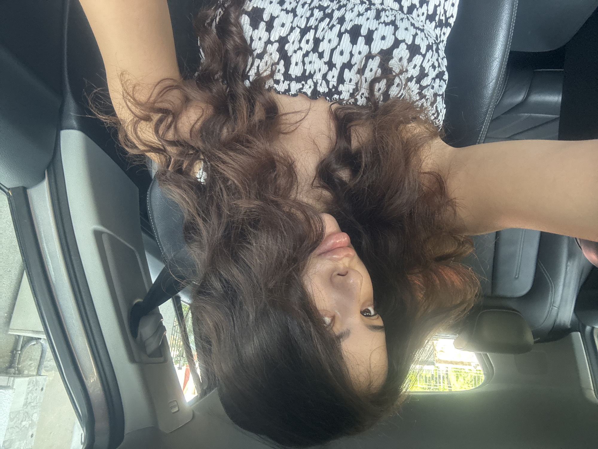 A young woman with long, dark, wavy hair is seated in the driver's seat of a car, holding a phone to take a selfie. She is wearing a strapless black and white floral print top. The interior of the car appears to be black leather. Outside the car, through the rear window, vegetation is visible, suggesting an outdoor location, possibly on the outskirts of a city. The scene is lit by natural daylight. The specific location in the city of Plovdiv, Bulgaria, cannot be determined from the visual information provided.