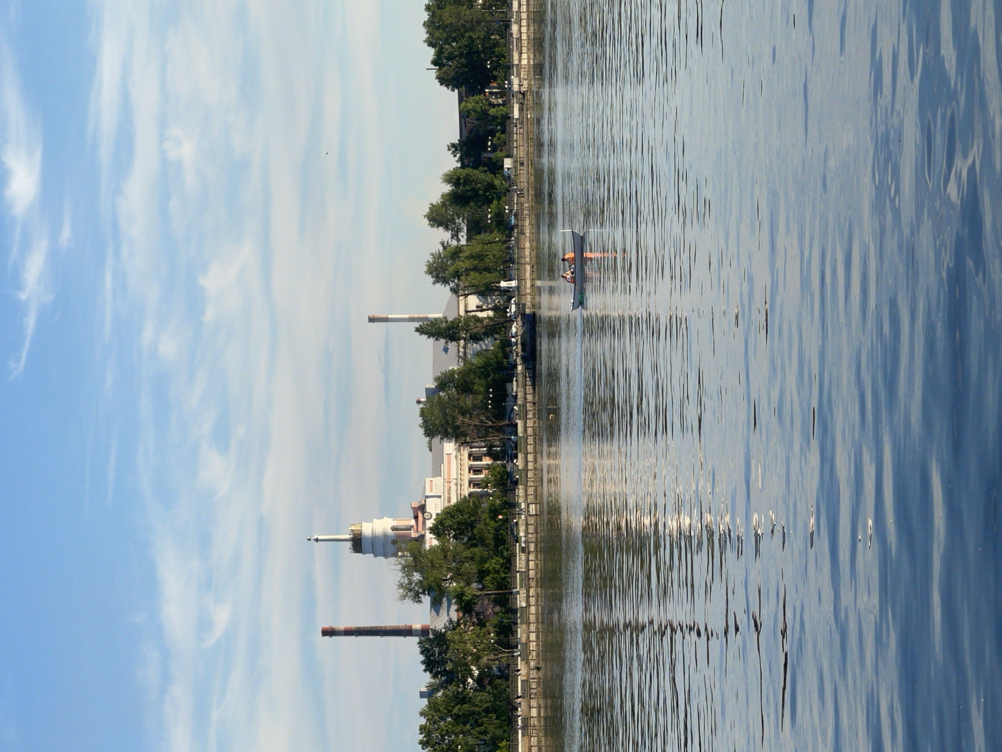 A distant view of a factory and a body of water is depicted. The factory has a tall red and white smokestack on the left and a white tower with a spire on the right. The building itself is white with pink accents and features columns and large windows. Trees and a sidewalk line the front of the factory. In the water, a small white boat with two people wearing orange vests is visible, rowing across the calm surface. The sky is blue with scattered white clouds. The overall scene suggests a peaceful day in an industrial area with recreational activity on the water.
