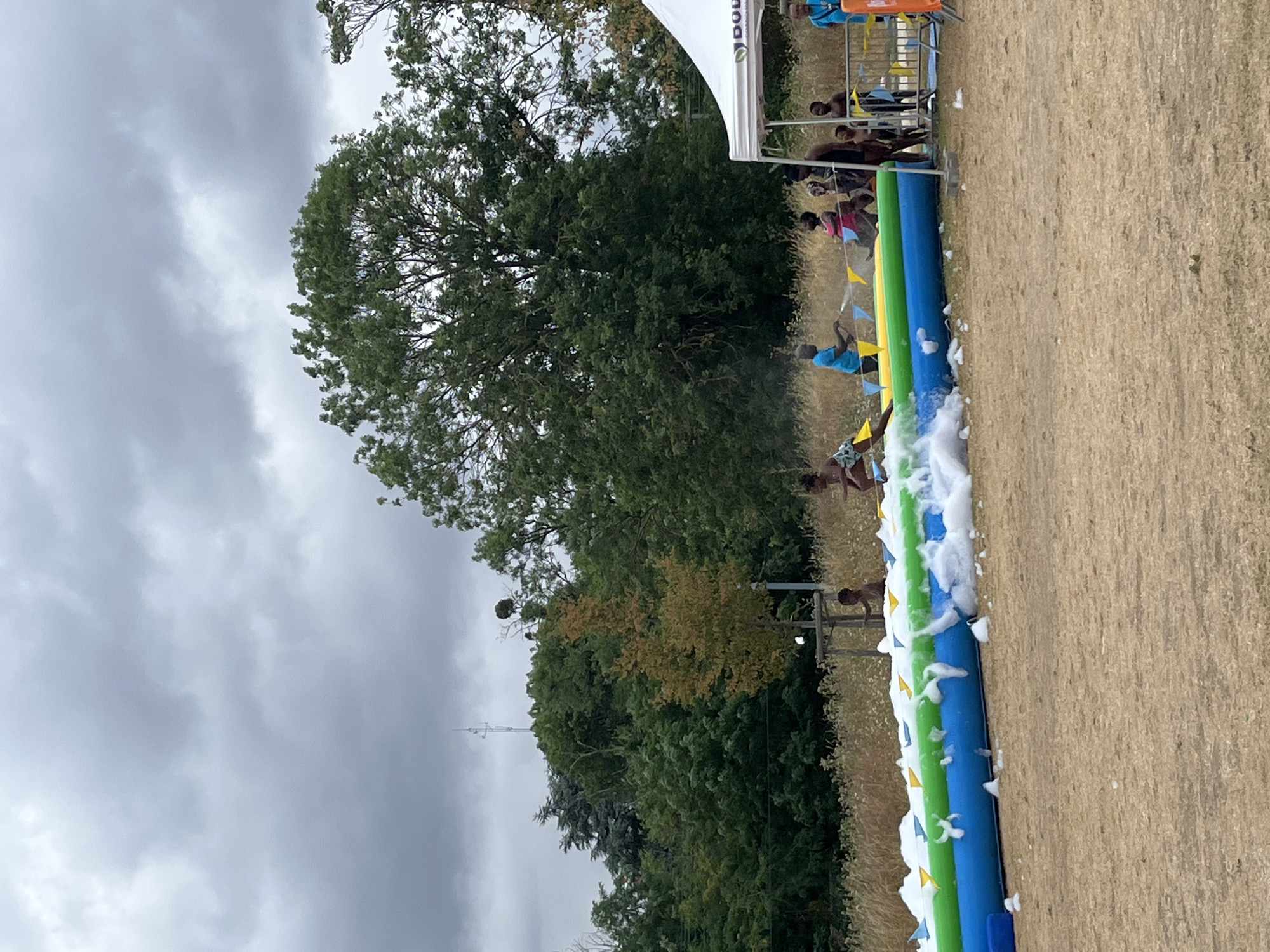 In Bobigny, France, on a cloudy day, a group of children and adults are gathered near an inflatable water slide covered in foam. The slide is long and multi-colored, with blue, green, and yellow sections. Some children are actively playing on the slide, sliding down with foamy water. Others are standing nearby, watching or preparing to participate. A white tent with branding is set up to the right of the slide, offering shade. The background consists of trees and dry grass, suggesting an outdoor recreational area. A tall communication tower is visible in the distance, over the trees. The ground is covered in dry, light-colored earth or sand.