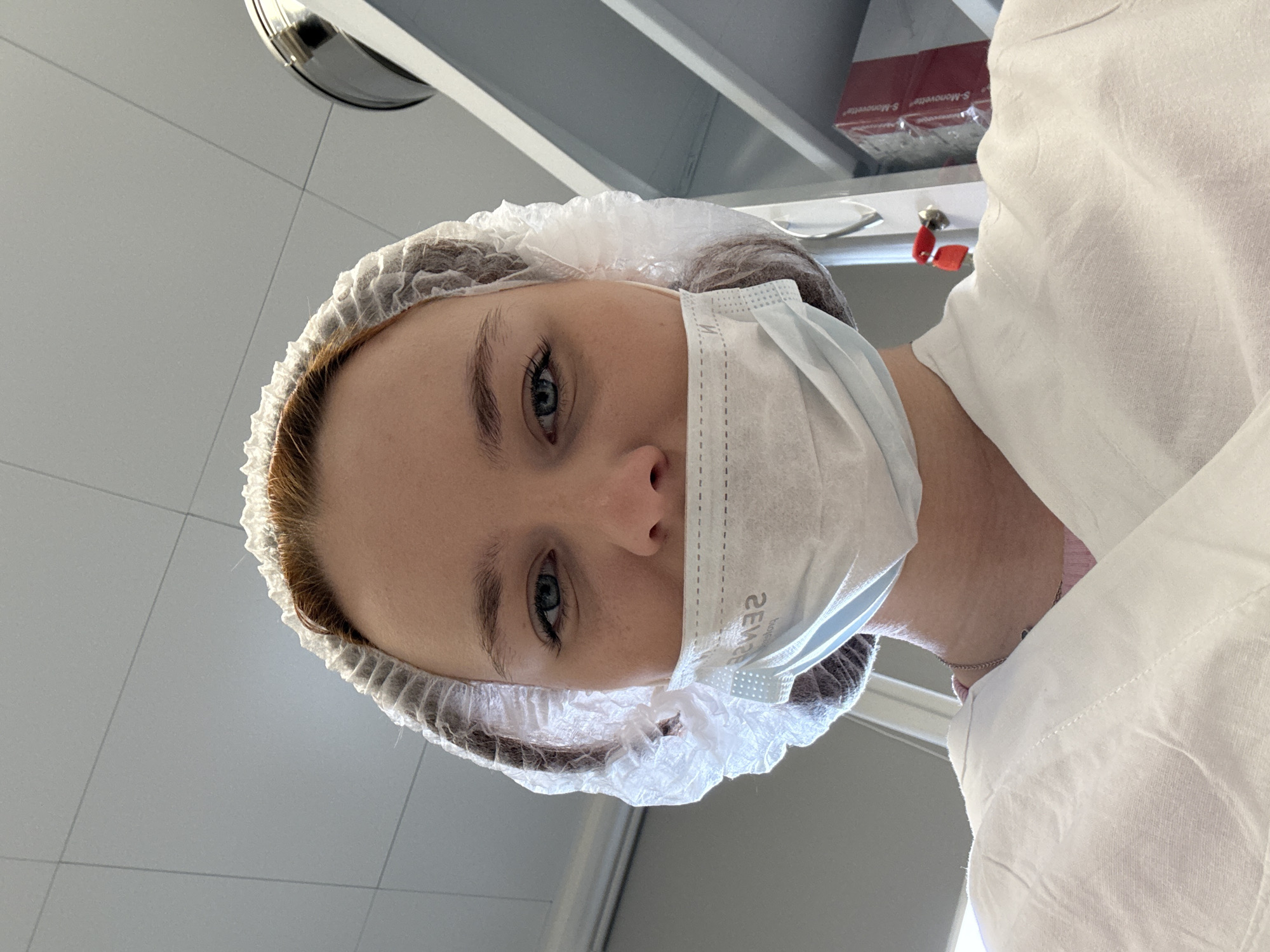 A woman wearing a white hairnet, a surgical mask, and a white gown looks down at the viewer. She is in a sterile environment, possibly a clinic or hospital room, with white tiled walls and medical equipment visible in the background. The location is unknown, and there is no information to suggest that it is in Saint Petersburg, Russia.