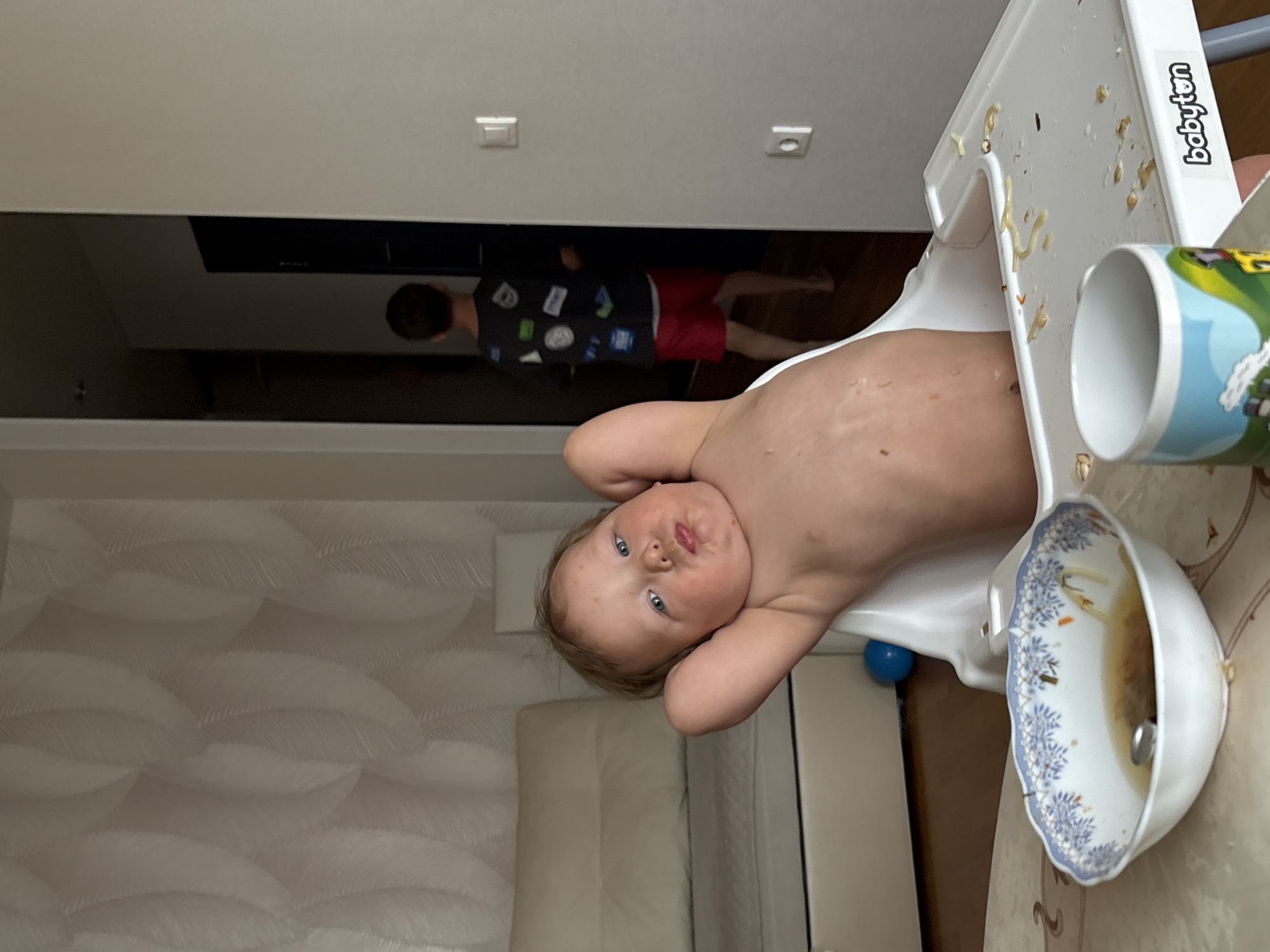 A child is in a high chair in a room in Chaikovsky, Russia. The child has blue eyes and appears to be shirtless. There is a bowl with food in it and a mug on the tray of the high chair. In the background, a second child is wearing a black shirt with colorful patches and red shorts. The room has white walls and a dark opening to another room. There are ceiling lights visible. The high chair is a "babyton" brand. The image is rotated.
