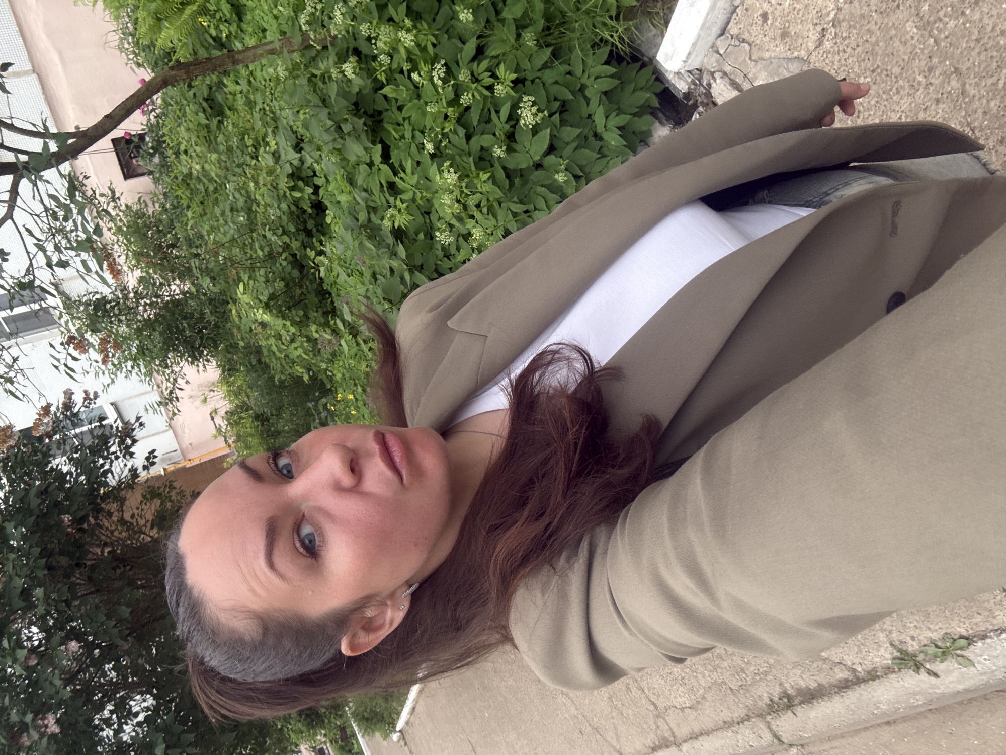 A medium-shot selfie captures a young woman outdoors in Chaikovsky, Russia. The woman, with long dark brown hair pulled back from her face, blue eyes, and a neutral expression, looks directly into the camera. She is wearing a white t-shirt under a loose-fitting khaki or olive-green blazer or jacket. Small earrings are visible on her ears.
The background is dominated by lush green foliage, including dense bushes and trees, suggesting a vibrant natural setting. Behind the greenery, parts of a light-colored building, possibly pale pink or beige, are visible, along with a partially obscured window. The ground beneath and to the side of the woman is a cracked concrete or paved surface, indicative of a sidewalk or pedestrian area, with a curb or low wall running along its edge. The lighting appears soft and diffused, suggesting it is daytime, possibly overcast or in a shaded area during a warm season like spring or summer. The woman is holding the camera herself, taking a close-up self-portrait. No text is visible in the image.