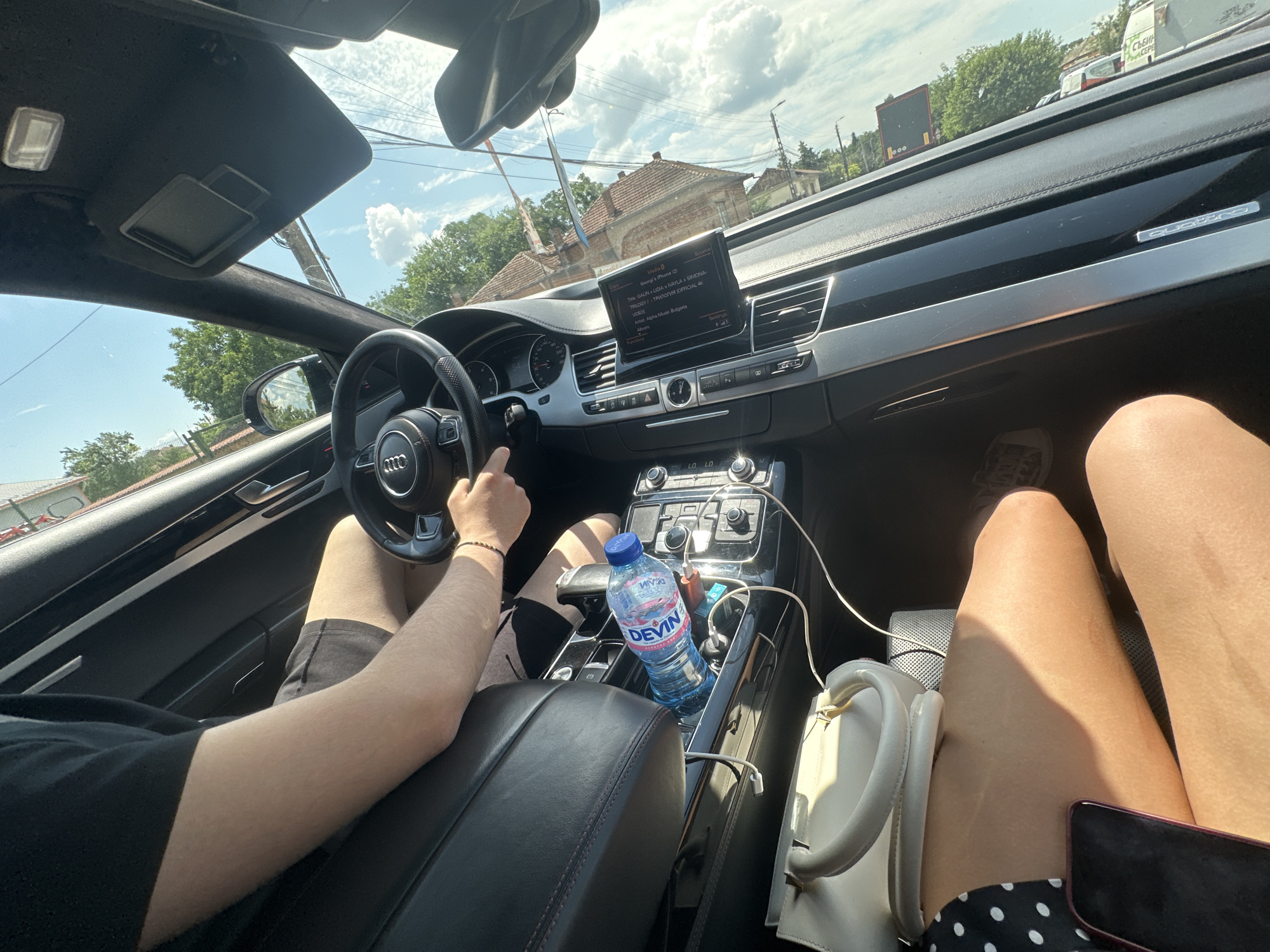 This media file captures an interior view of a car, likely an Audi, as it travels along a road in Polikraishte, Bulgaria. The perspective is from the passenger seat, looking towards the driver and the car's dashboard.
The primary subjects are two people: the driver, whose hands are on the steering wheel and who is wearing black shorts and a black t-shirt, and a passenger, whose legs and part of a white handbag are visible in the foreground, wearing a black and white polka dot skirt. The car's interior features black leather seats, a modern dashboard with a central display screen, and a center console.
The central display shows information from a connected iPhone, with song titles like "GALIN x LIDIA x IVAYLA x SIMONA - TRILOGY" and artist/album details. This indicates that music is playing. A bottle of DEVIN brand water is prominently placed in a cup holder on the center console, along with charging cables.
The scene is set during the daytime, as indicated by the bright sunlight illuminating the interior and casting shadows. The weather appears to be clear and sunny, with a blue sky and scattered clouds visible through the windshield and sunroof. Outside the car, residential buildings with red-tiled roofs and trees are visible, consistent with a suburban or rural setting. The presence of a sign with Cyrillic text, "СЪБИН ACEPE", further confirms the Bulgarian location. The overall mood appears relaxed, suggesting a casual drive.