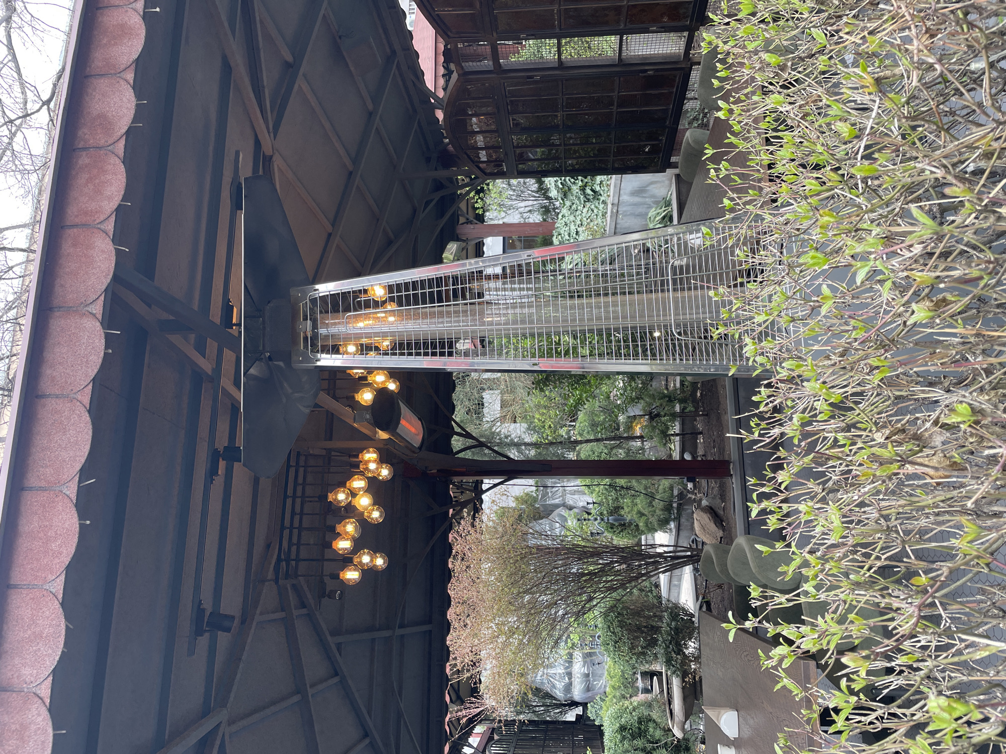 The image depicts an outdoor dining area, likely a patio or garden extension of a restaurant or cafe. The location appears to be in Yerevan, Armenia, based on architectural styles and vegetation.
Overhead, a wooden structure with a tiled roof provides partial cover. A metal-caged heater stands tall within the structure, suggesting a cooler climate. A string of decorative lights hangs nearby.
Below, trees and greenery are visible. Garden features like a white, semi-transparent covering can be seen, adding to the outdoor aesthetic.
In the foreground, a bush or shrub with budding leaves is the closest element to the viewer. A table with chairs is partially visible, as well as several lounge chairs, and suggesting seating for customers.