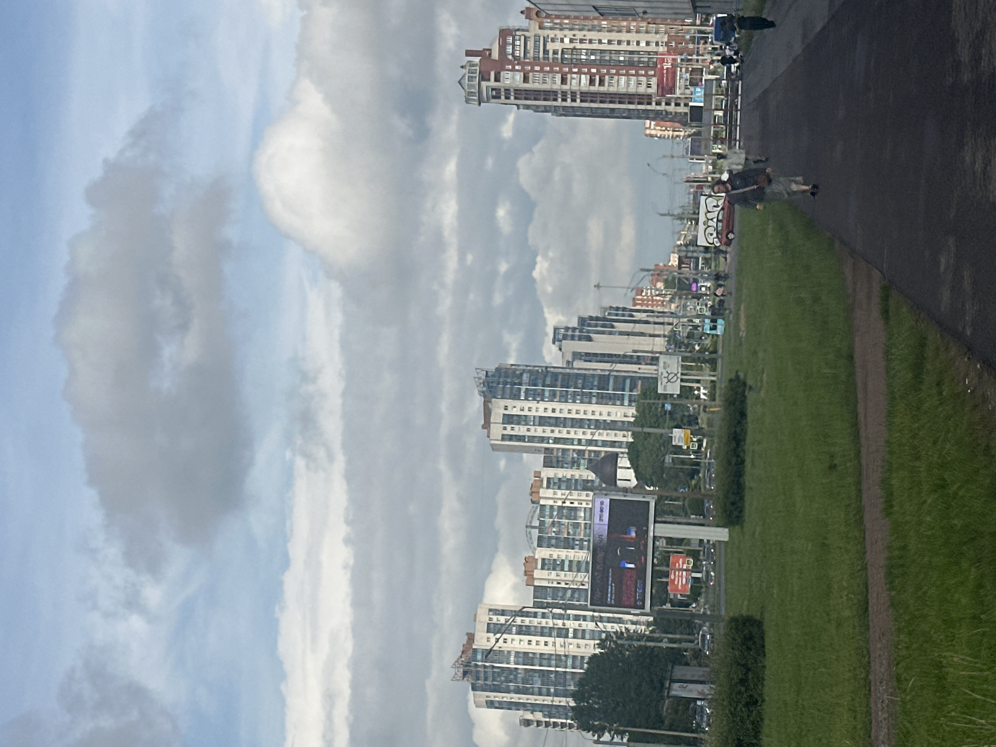 The image shows a street scene in an urban environment with several tall residential buildings. The sky is cloudy, with patches of blue visible. In the foreground, there is a grassy area with a paved path running alongside it. Several people are walking on the path. In the background, there are cars, traffic lights, and advertisements. The buildings are modern, with many windows and balconies. One building on the right is particularly tall and has a distinctive red and white facade. Another building on the left appears to be a cluster of connected towers.