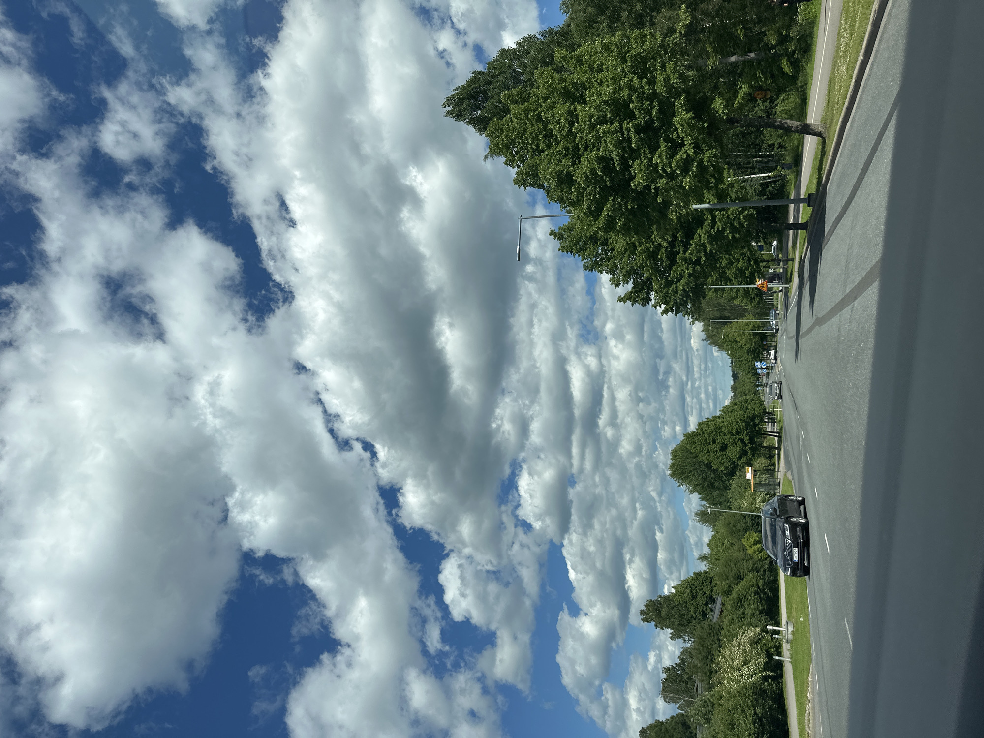 The image depicts a scenic view from a vehicle traveling along a road. The scene is dominated by a vast expanse of blue sky filled with dramatic, layered white clouds, suggesting a partly cloudy day with dynamic weather. Tall, lush green trees line the right side of the road, creating a vibrant canopy and a sense of natural beauty. The road itself appears to be paved, with a distinct lane marking visible, indicating it is a well-maintained thoroughfare.
A dark-colored car is the only prominent man-made object visible on the road, traveling in the same direction as the viewer. Its presence suggests ongoing transit. The time of day appears to be daytime, likely late morning or afternoon, given the bright sunlight casting shadows and illuminating the clouds.
Notable details include streetlights positioned along the roadside, hinting at an urban or suburban setting where such infrastructure is common. The overall impression is one of a pleasant journey through a green landscape under a striking sky. There is no visible text in the image.