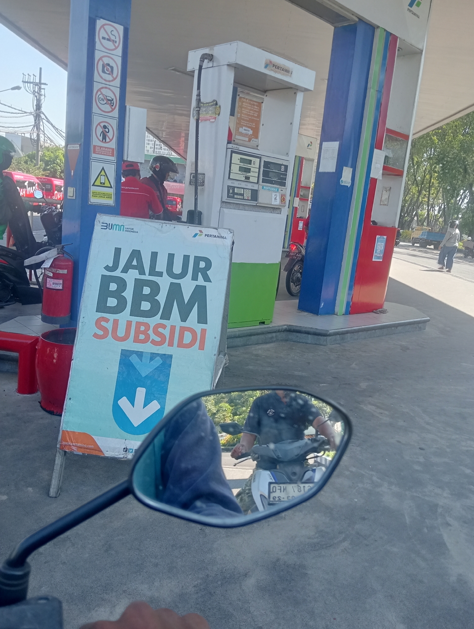 The image captures a scene at a Pertamina gas station in Surabaya, Indonesia, likely on a bright, sunny day. In the foreground, a motorcycle's rearview mirror reflects the rider, who is wearing a dark shirt and a helmet. The rider's hand is visible on the handlebar.
Dominating the mid-ground is a large, light blue sign with prominent white text that reads "JALUR BBM SUBSIDI," which translates to "Subsidized Fuel Lane." Below this text is a large, downward-pointing blue arrow. This sign is positioned in front of a Pertamina gas pump, which is painted with the brand's signature blue, red, and green colors. A red fire extinguisher and a red trash bin are also visible near the sign.
In the background, several other Pertamina gas pumps are visible, along with people and vehicles. One person wearing a red uniform and a helmet is attending to a customer at a pump. Other individuals and a truck are seen further in the background, suggesting a typical day at a busy gas station. The environment appears to be an open-air space, with trees and power lines visible in the distance, indicating an urban setting. The pavement is a light gray concrete.