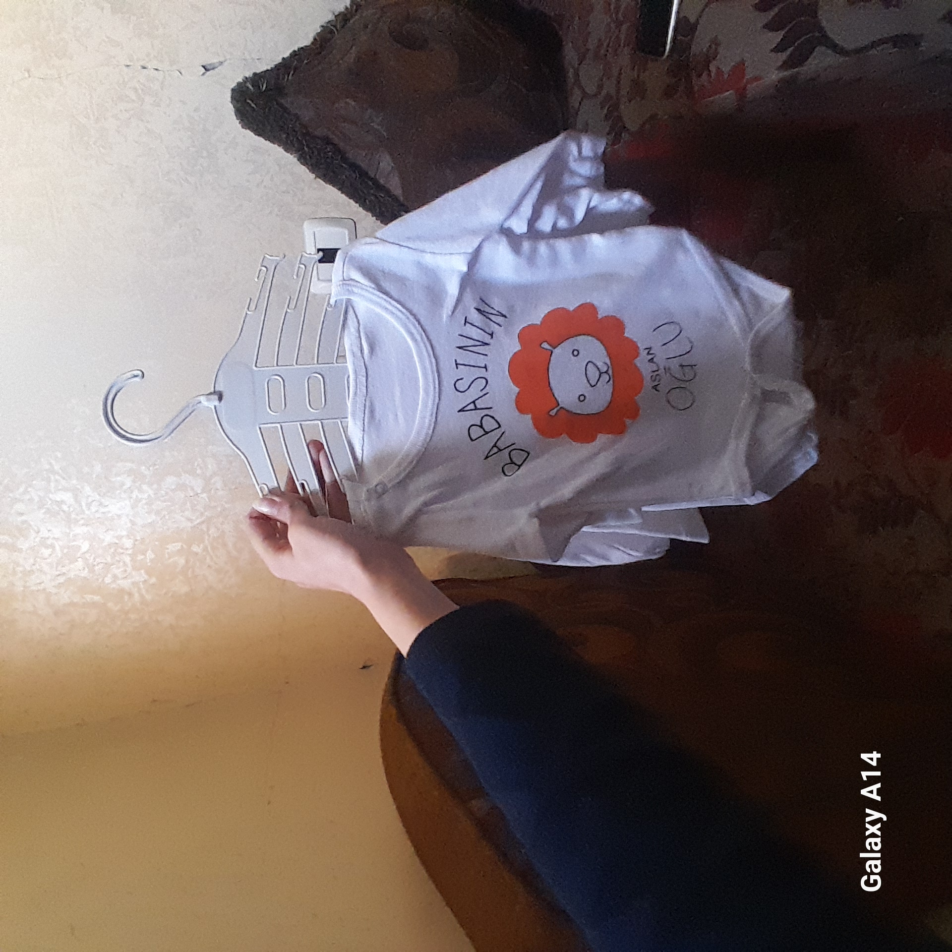 A hand holds a white hanger with a white t-shirt on it. The t-shirt has an orange lion cartoon on it with text in an unknown language. A pillow rests behind the hanger and t-shirt on a brown surface. A partial view of a table and arm are visible in the lower right corner. The image includes text, "Galaxy A14" in the lower right corner. The location is Unknown, Unknown.