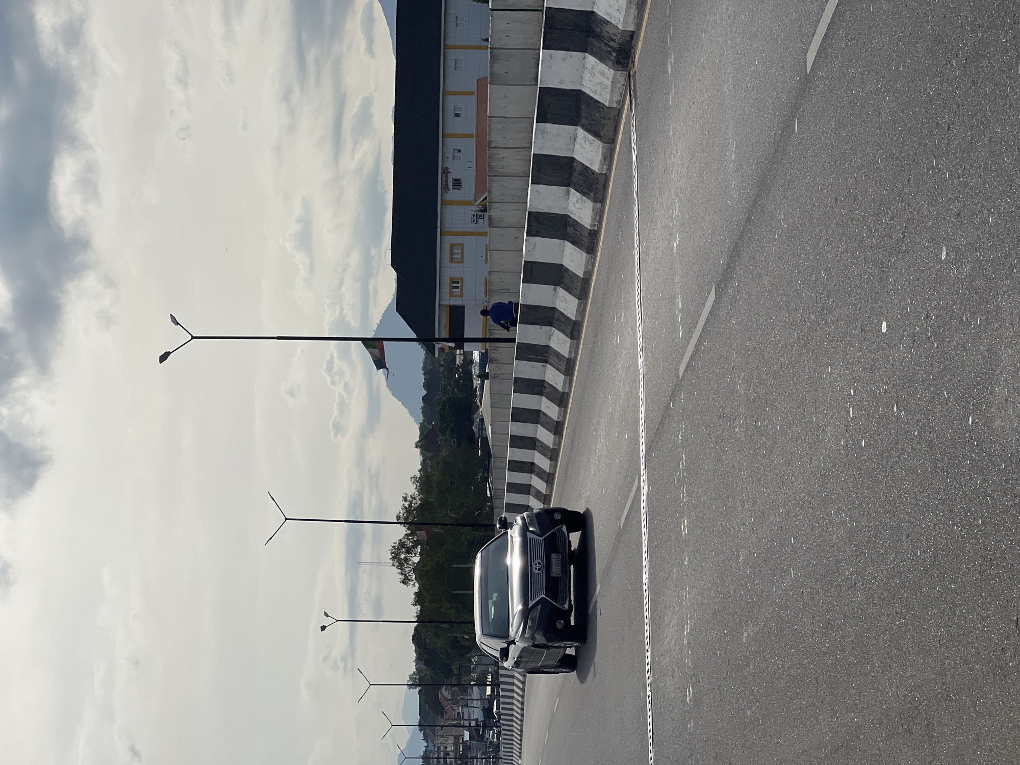 The image captures a view of a highway overpass in Jalingo, Nigeria. A black sedan, possibly a Lexus, is driving on the overpass, its headlights reflecting the cloudy sky. The highway is lined with a concrete barrier painted with alternating black and white stripes, a common safety feature. In the background, a building with a dark roof and yellow accents stands out, and a Nigerian flag flutters on a pole. A person on a motorcycle is visible behind the barrier, near the building. Distant hills form the backdrop under a sky filled with a mix of bright clouds and some patches of blue. The road surface shows lane markings and a metal expansion joint.