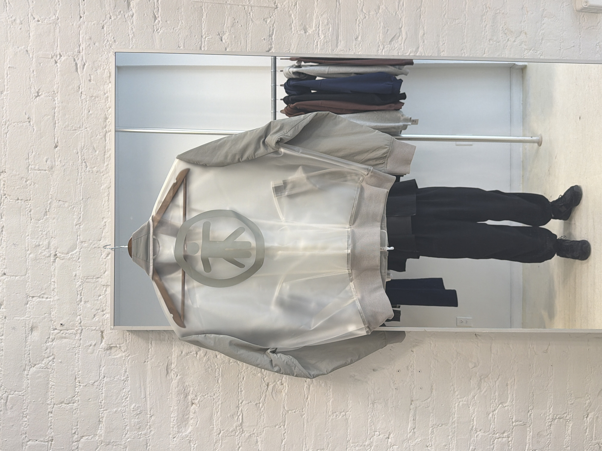 A horizontally oriented, medium-shot captures a reflection of clothing and a person within a shop setting. A bomber jacket made of translucent material with a gray trim and a logo in the center hangs on a wooden hanger. In the reflection, a person in black pants and black sneakers is visible, with clothing stacked on a shelf above them. The reflection appears to be of a shop in a city. The background is a white brick wall. Based on the clothing and overall aesthetic, the location could be a boutique clothing store in New York City, United States.
