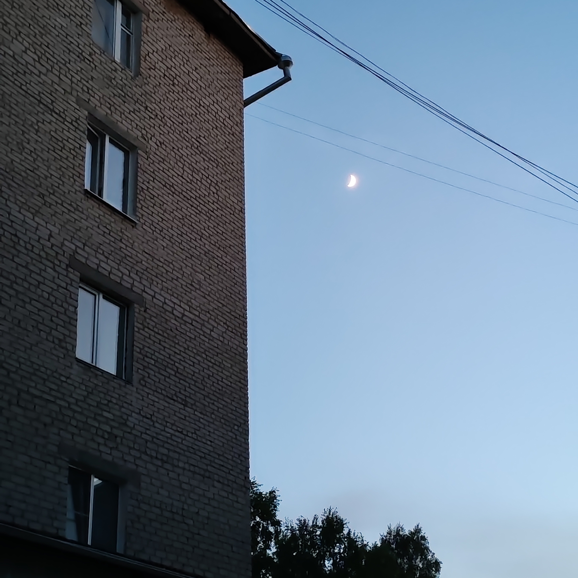 A brick building occupies the left side of the frame, featuring multiple windows. Electrical wires are visible against the sky, which displays a crescent moon. The location is likely Chaikovsky, Russia, based on associated information. Trees are also visible at the bottom of the image.