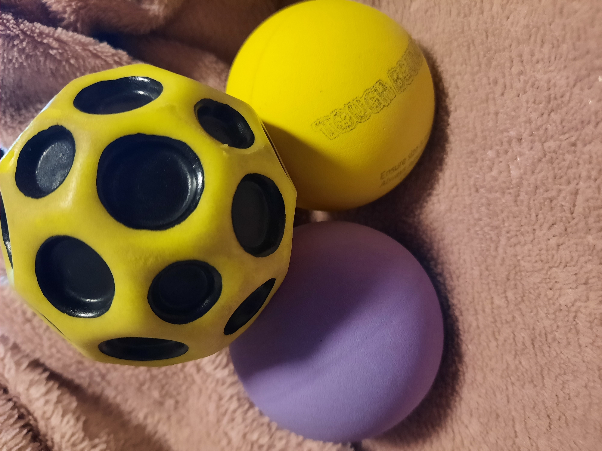 The image shows three balls stacked on a textured surface, which appears to be a blanket. The top ball is yellow and has a unique geometric shape with several black circular indentations. Below this, to the left, is a solid purple ball. To the right of the purple ball and slightly behind it is a solid yellow ball. This yellow ball has some faint writing or logos on its surface. The background is blurred and consists of the same textured material as the surface the balls are resting on. There is no indication of the location being in Rosenheim, Germany.
