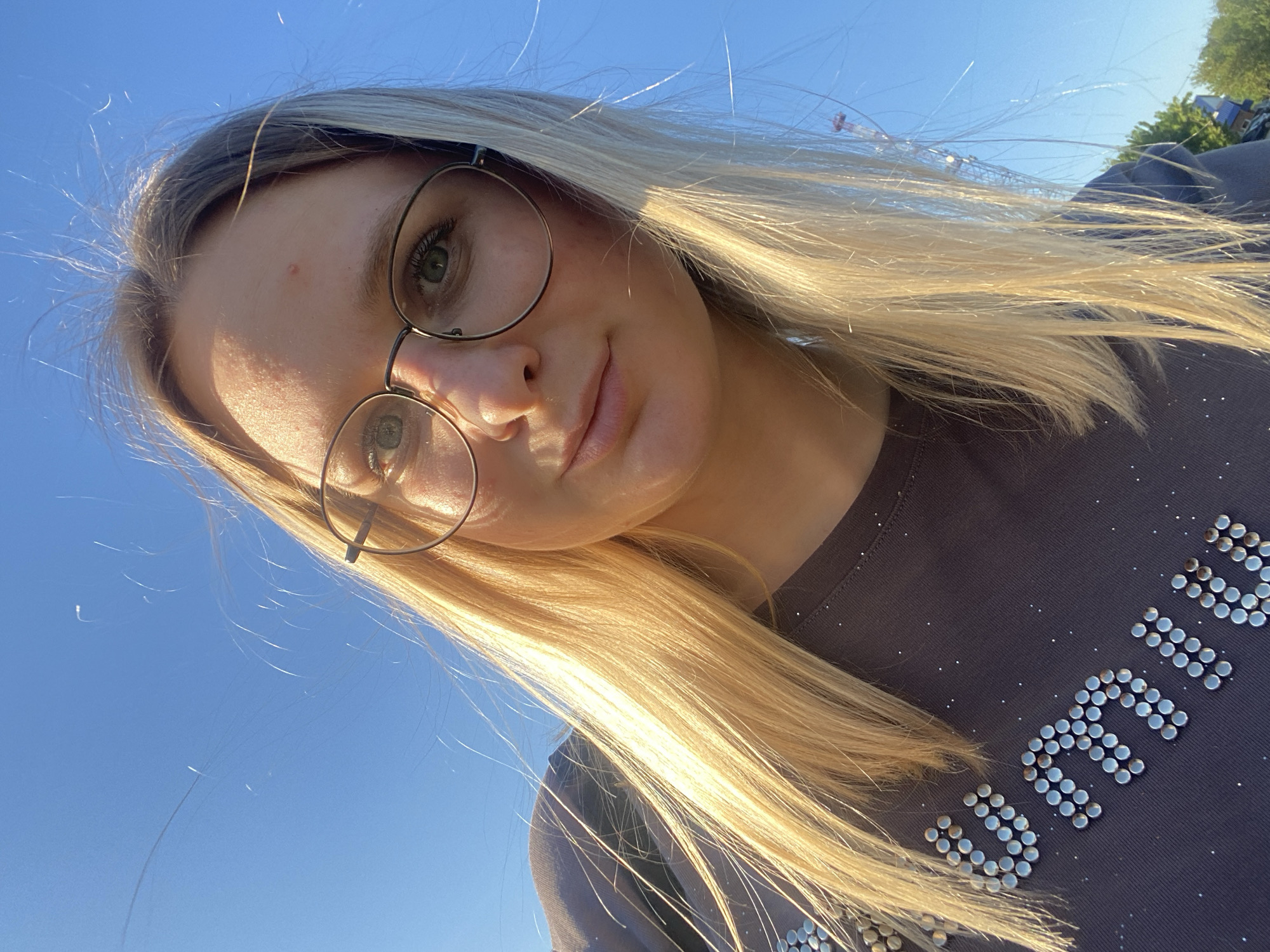 A low-angle, medium shot captures a young woman with fair skin and long, blonde hair, wearing glasses. She is positioned against a backdrop of a clear, blue sky. The woman’s face is slightly angled towards the left. She is wearing a dark t-shirt with silver studs spelling out a word. A portion of a building or tree is visible in the upper right corner, suggesting an outdoor location.