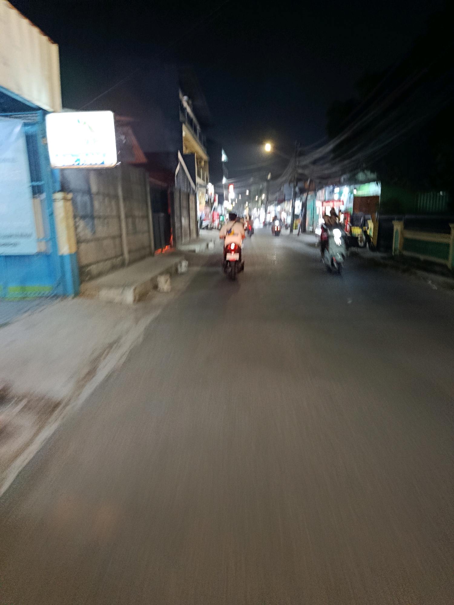The image captures a night scene on a paved road in the city of Bekasi, Indonesia. The perspective suggests the photo was taken from a moving vehicle, likely a motorcycle, given the motion blur. The road is flanked by buildings and structures, some of which are illuminated by artificial lights, indicating commercial activity or residences. Several motorcycles are visible on the road, some with riders, suggesting traffic and transportation are active. Streetlights and building lights cast a dim glow on the surroundings, creating a somewhat hazy atmosphere typical of urban night environments. The overall impression is one of a busy street in Bekasi at night, with the blur of movement adding a dynamic element to the scene.
