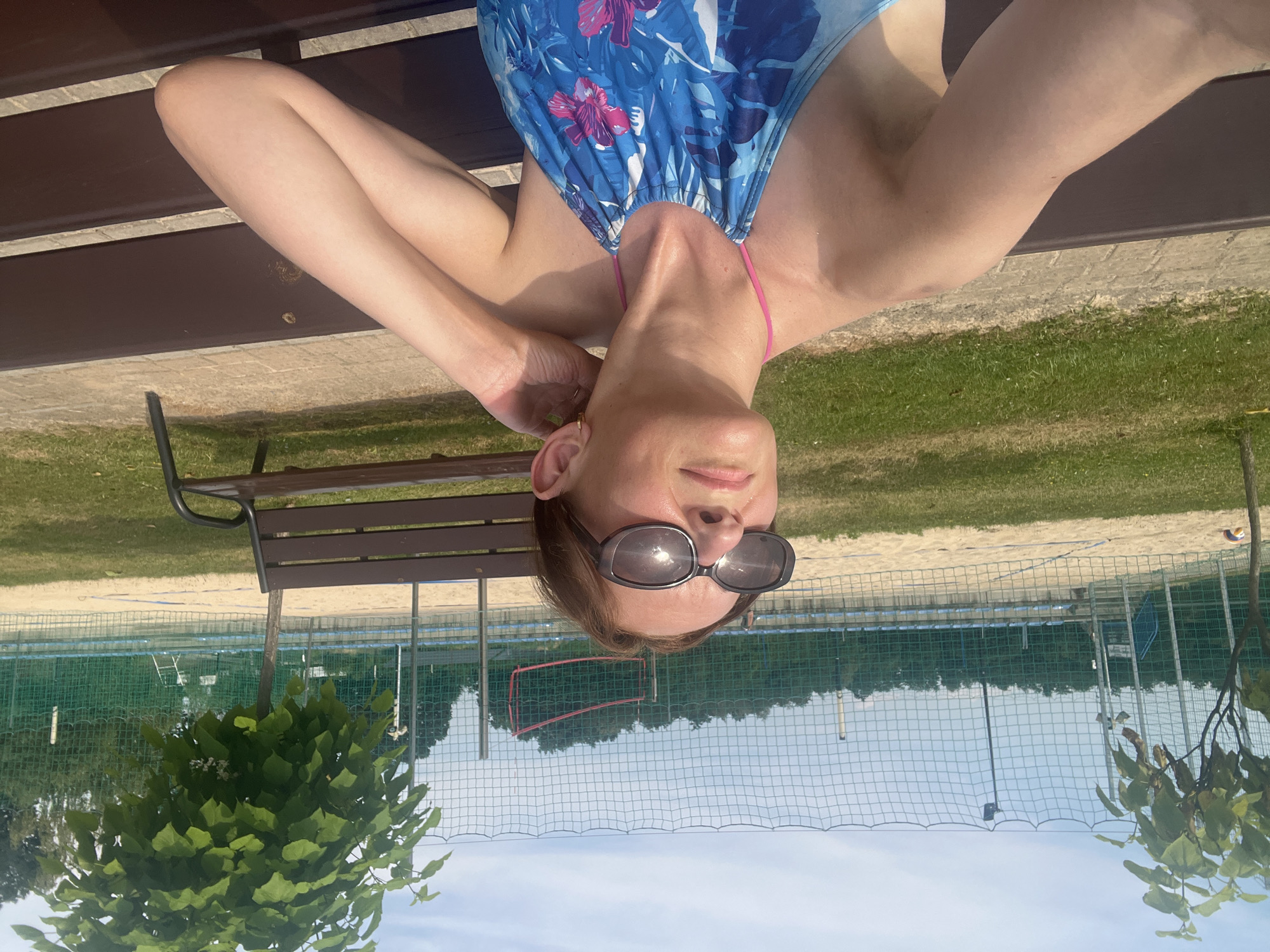 This is a horizontal, upside-down selfie of a person lying on their back on a wooden bench. The person is wearing a blue and pink floral swimsuit and sunglasses. Their arms are behind their head, and their face is looking up. The background shows a grassy area, a sand volleyball court with a net, and trees. The sky is visible in the upper part of the image, with some clouds. The lighting suggests it is daytime, possibly sunny. The overall mood appears relaxed and casual, typical of a person enjoying leisure time outdoors.