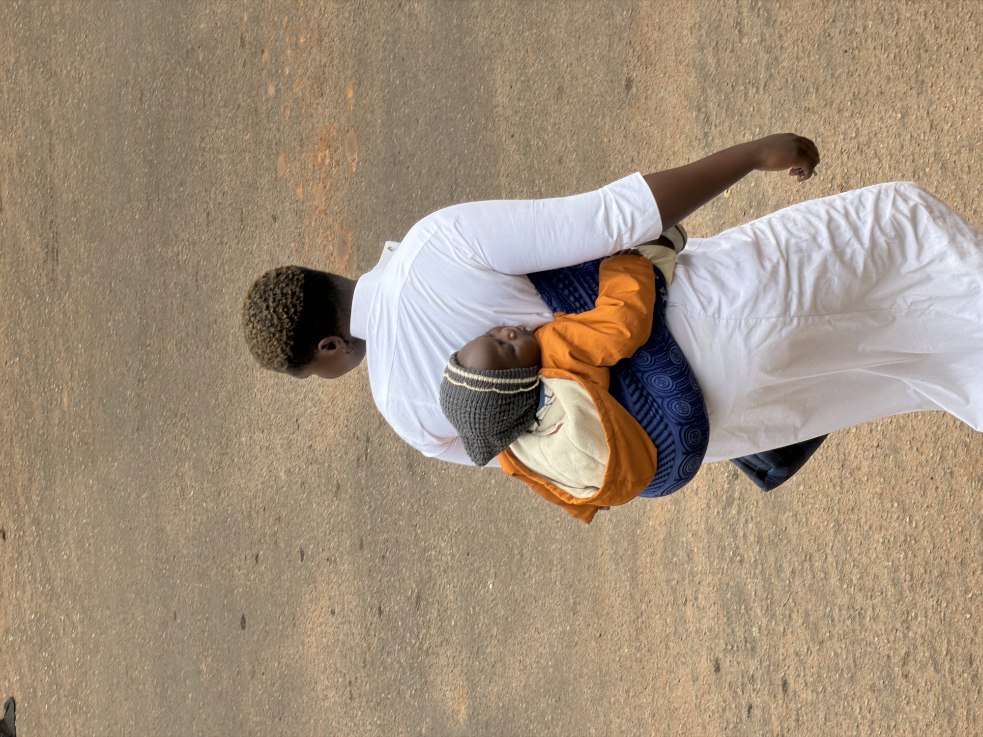 A person, viewed from behind, is walking on a paved surface. They are carrying a baby on their back in a blue and white fabric sling. The baby is wearing an orange jacket and a grey and white striped hat. The person is wearing a white garment. The background is a textured, light brown surface, possibly a road or path. The scene appears to be in Bukuru, Nigeria.