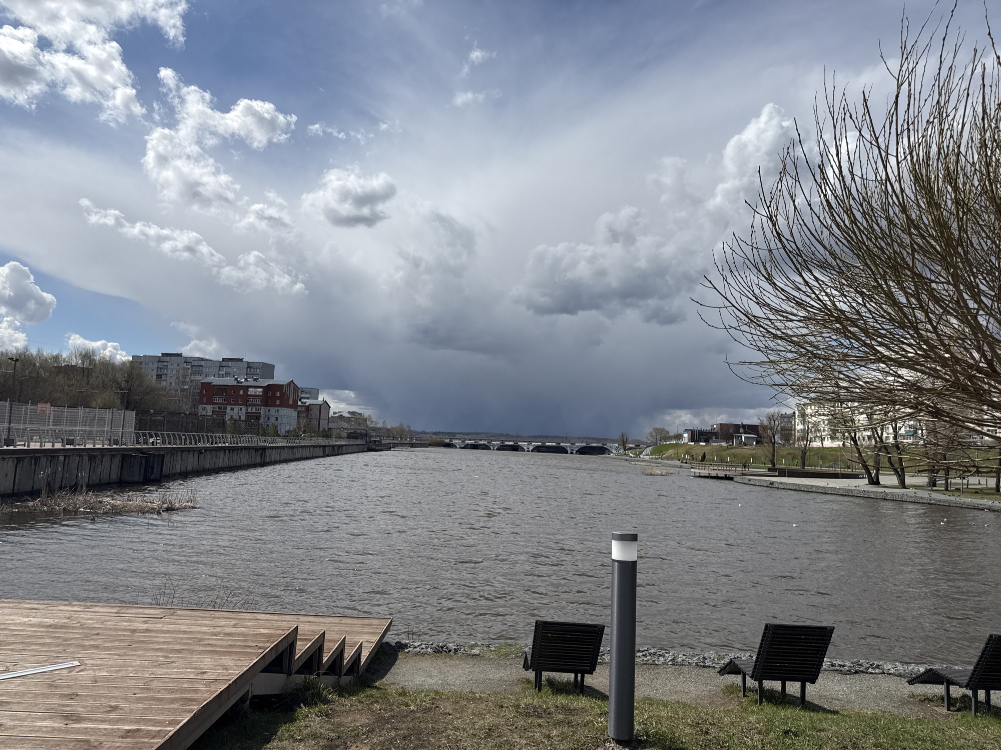 This media file captures a wide, scenic view of a river or large canal in Votkinsk, Russia, under a dramatic sky. The image is taken from the right bank, looking across the water towards the opposite side and downriver.
In the foreground on the left, a wooden deck with stepped sections extends towards the water. Beyond it, a concrete embankment with a metal railing runs along the left bank, leading to urban buildings, some with red roofs, in the background. Sparse reeds are visible in the water near the wooden structure.
On the right side of the foreground, bare tree branches frame the upper right corner, indicating early spring or late autumn. The ground is covered with sparse grass and gravel, where three dark, slatted benches and a modern grey lamp post stand near the water's edge.
The wide expanse of water appears choppy with ripples, suggesting wind, and is a dark grey color. In the distance downriver, a multi-arched bridge spans the waterway, with more urban development and green spaces visible on the banks beyond it.
The sky is a prominent feature, showcasing a clear contrast: bright blue sky with fluffy white clouds on the left gives way to heavy, dark grey storm clouds dominating the center and right, hinting at impending rain or a strong weather front. The overall impression is daytime, possibly a moody afternoon. The scene is devoid of people, presenting a tranquil yet atmospheric waterfront landscape.