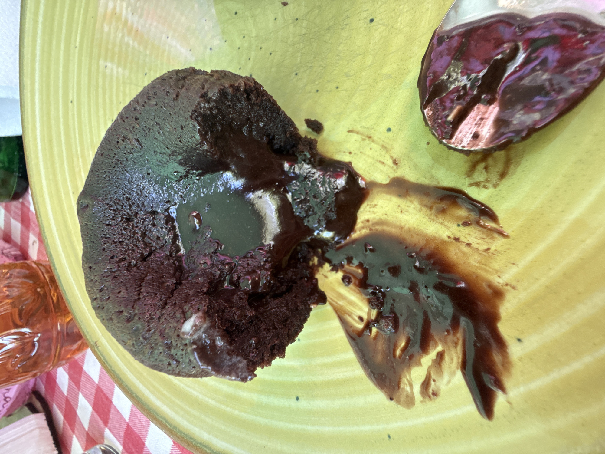 A partially eaten chocolate lava cake sits on a yellow plate alongside a spoon with remnants of the cake. The plate rests on a table with a red and white checkered tablecloth. A jar is partially visible in the background. The location is not discernible as Bliznatsi, Bulgaria based on the objects in the photo.