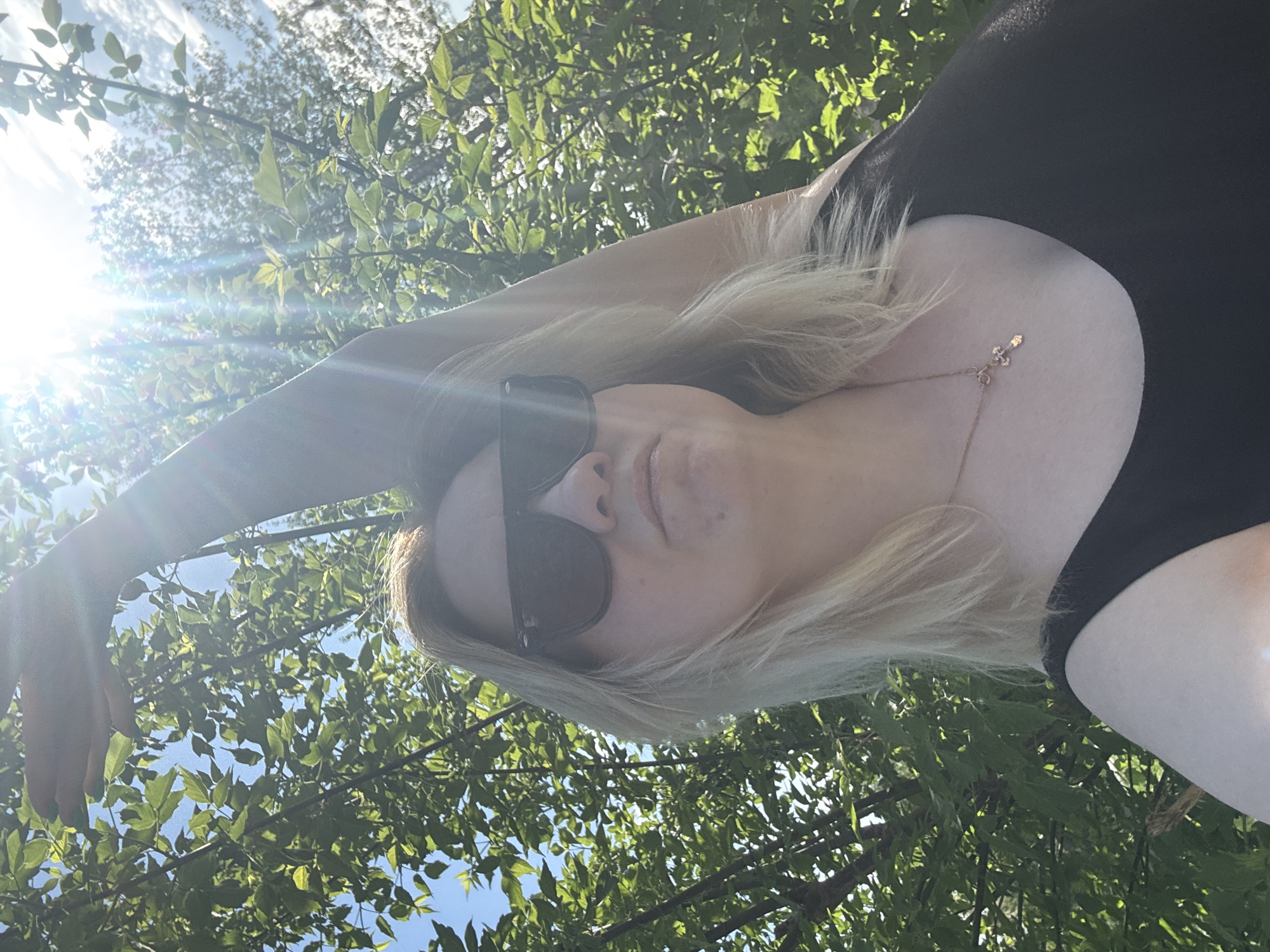 A woman with blonde hair and wearing sunglasses is looking up at the camera from a low angle. She is wearing a black top and a delicate gold necklace. The background shows a tree canopy with green leaves, with glimpses of a blue sky and bright sunlight. The location cannot be confirmed as Chaikovsky, Russia from the provided image.