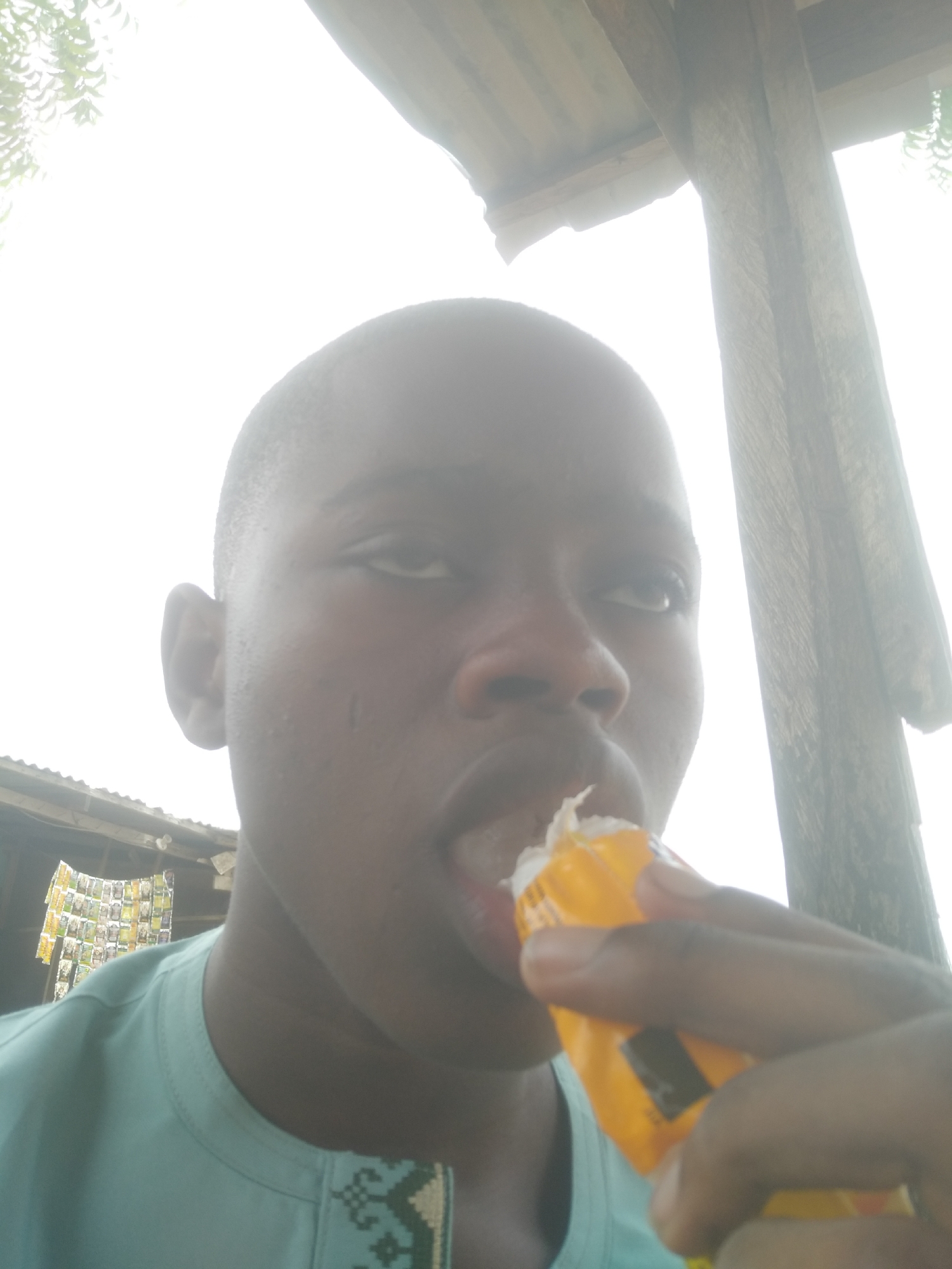 A person is eating an item from a yellow package. The environment includes a wooden structure overhead and another structure in the background, indicating an outdoor setting in an undefined urban area.