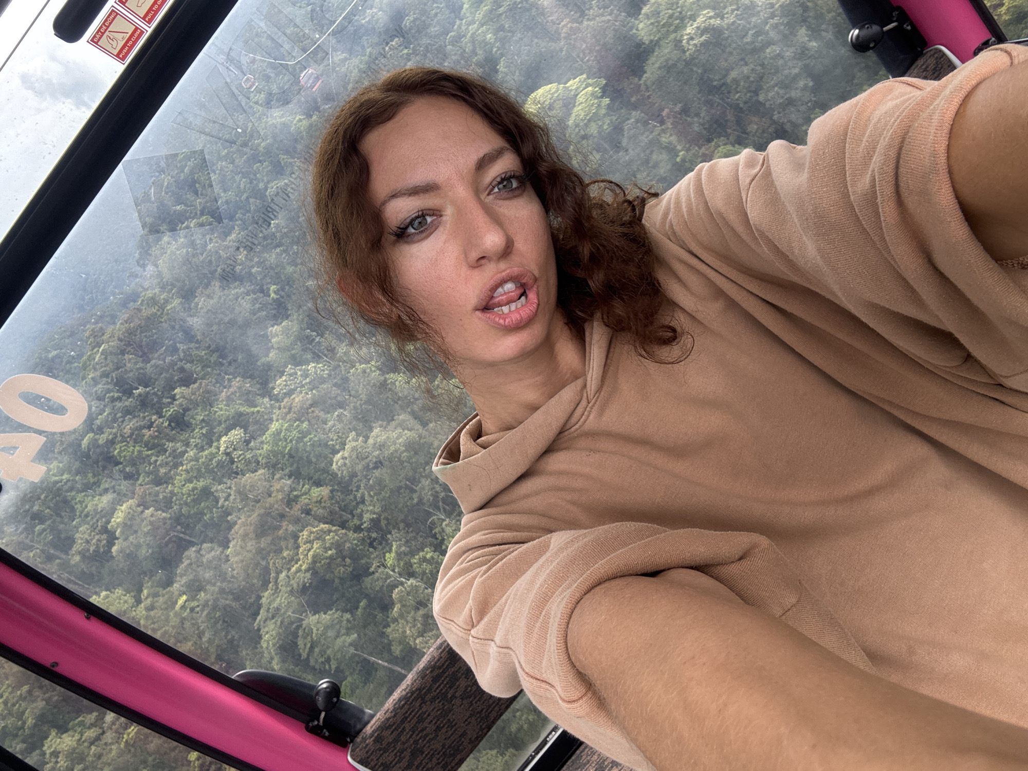 A selfie captured inside a cable car cabin shows a woman with long, curly brown hair and green eyes, wearing a light brown hooded sweatshirt. She holds the camera at an upward angle, looking directly at it with a playful expression, her tongue slightly protruding.
Behind her, through the clear window of the cable car, is a panoramic view of a dense, green, forested mountain landscape, appearing somewhat hazy or misty in the distance. The interior of the cable car features bright pink structural elements, a wooden-textured handrail, and safety warning stickers on the glass.
The activity taking place is the woman taking a selfie while riding in the cable car. The time of day is daytime, with soft, natural light, suggesting overcast or misty weather conditions over the mountains. The location is consistent with a mountainous region around Đà Nẵng, Vietnam, confirmed by the user-provided context and the presence of Vietnamese text.
Visible text includes a large number "40" on the window. Safety stickers bear English text "PULL TO CLOSE" and "PULL TO OPEN," alongside Vietnamese text "DẬY ĐỀ DÓNG." Faintly reflected on the glass is additional Vietnamese text, "VỊ TRÍ TRIỆT ĐỘ." The woman's expression conveys a lighthearted and playful mood.