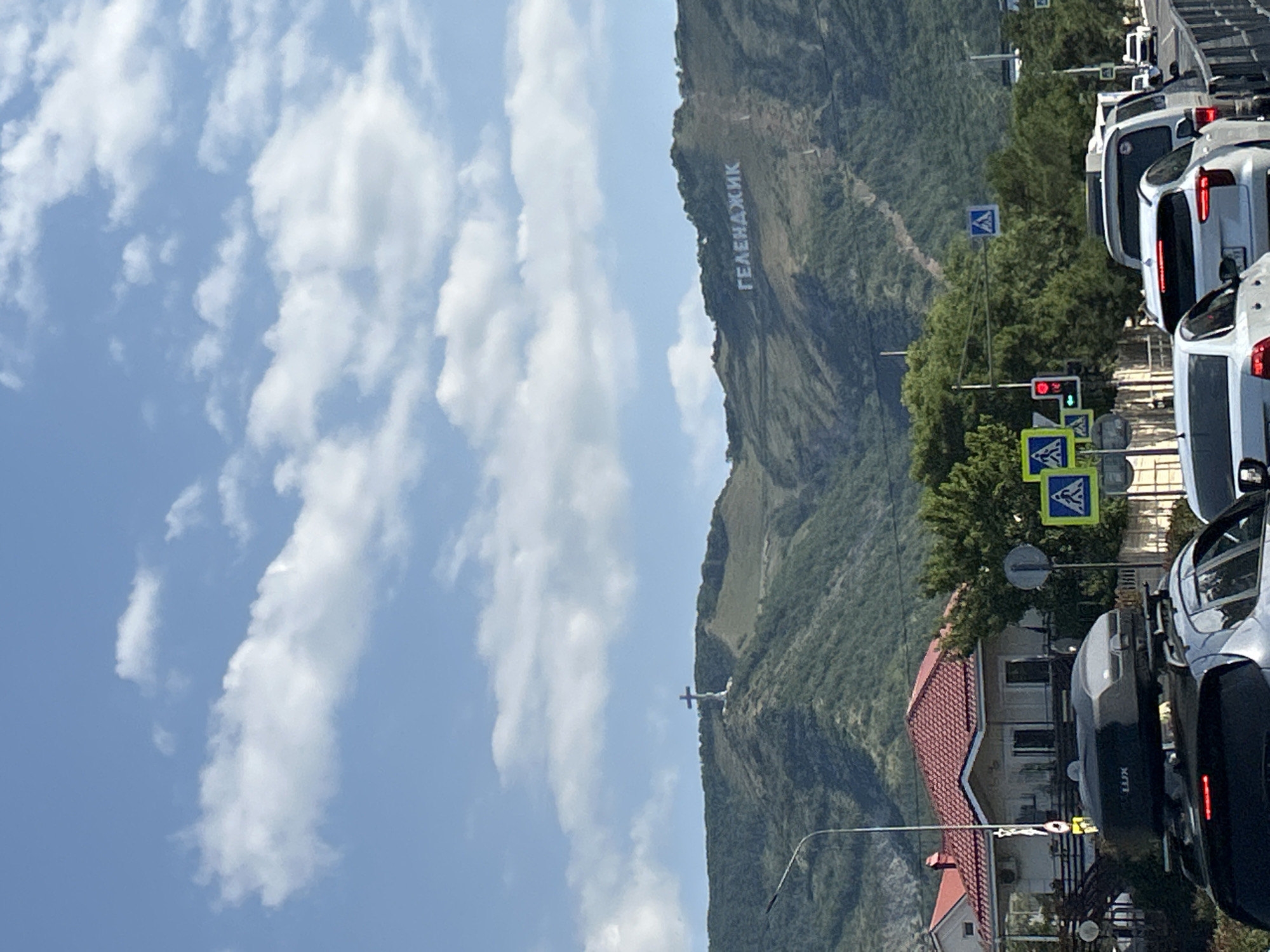 The image shows a view from a street with traffic, looking towards a large mountain range under a partly cloudy blue sky. On the mountain range, there is a large white cross on the left peak and white lettering spelling "ГЕЛЕНДЖИК" (Gelendzhik) on the right peak. The foreground shows a traffic jam with several cars. One car in the foreground has a black roof box. To the left of the road, there are some houses with red roofs, trees, and streetlights. Traffic lights and pedestrian crossing signs are visible. The overall scene depicts a sunny day in Gelendzhik, Russia, with the city's name prominently displayed on the mountainside and a significant cross as a landmark.