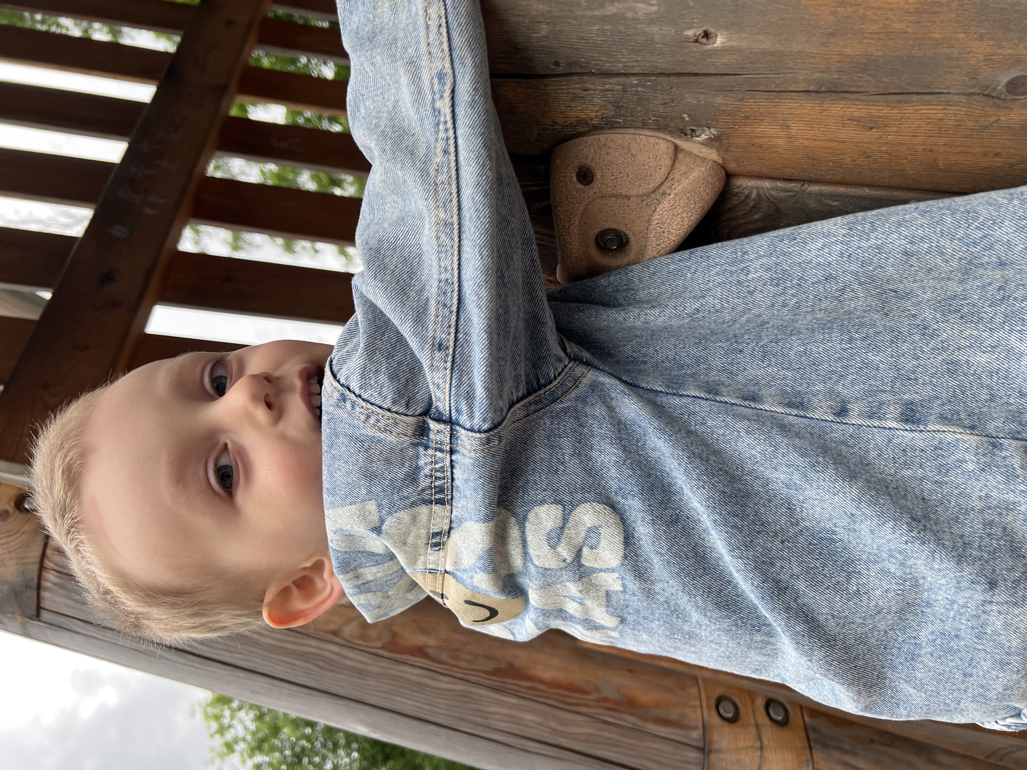 A child with blond hair hangs upside down on a wooden climbing structure. He is wearing a denim jacket. The climbing structure appears to be located outdoors, possibly in a park or playground. The precise location within Mytishchi, Russia, is indeterminable from the image.