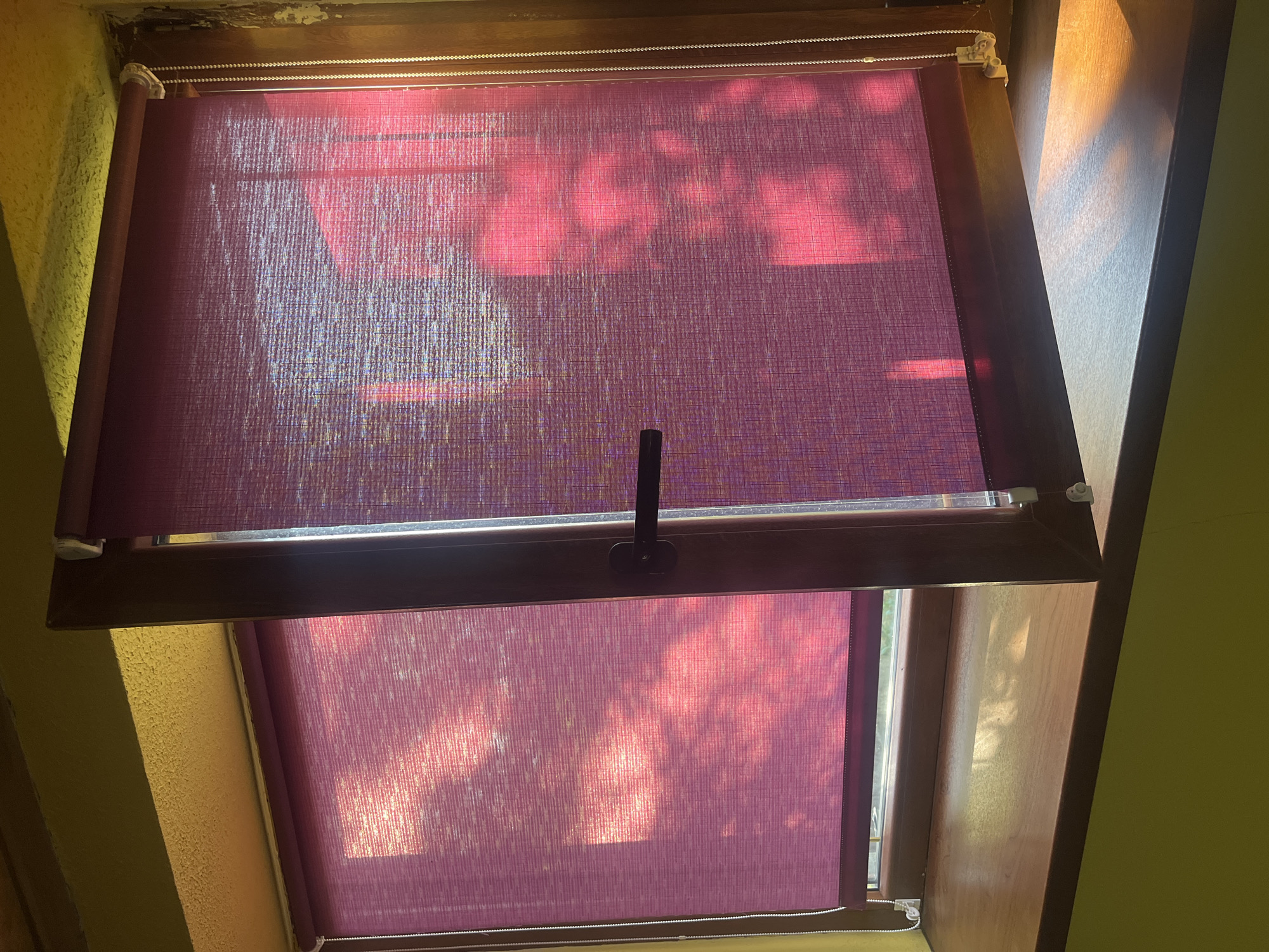 The image shows a close-up view of a windowsill with two purple roller blinds, one partially open. The blinds are made of a textured fabric and are illuminated by sunlight, which creates a dappled shadow effect from unseen foliage outside. The windowsill itself is made of wood and appears to be a light brown color. The wall to the left of the window is painted a bright yellow. The window frame is a dark brown color. There is a black handle on the partially open blind. The scene is indoors, looking out through a window. The location is identified as Tarnivtsi, Ukraine, though there are no specific geographical indicators within the image itself to confirm this. There are no people or animals present in the image. The overall impression is one of natural light filtering into a room.