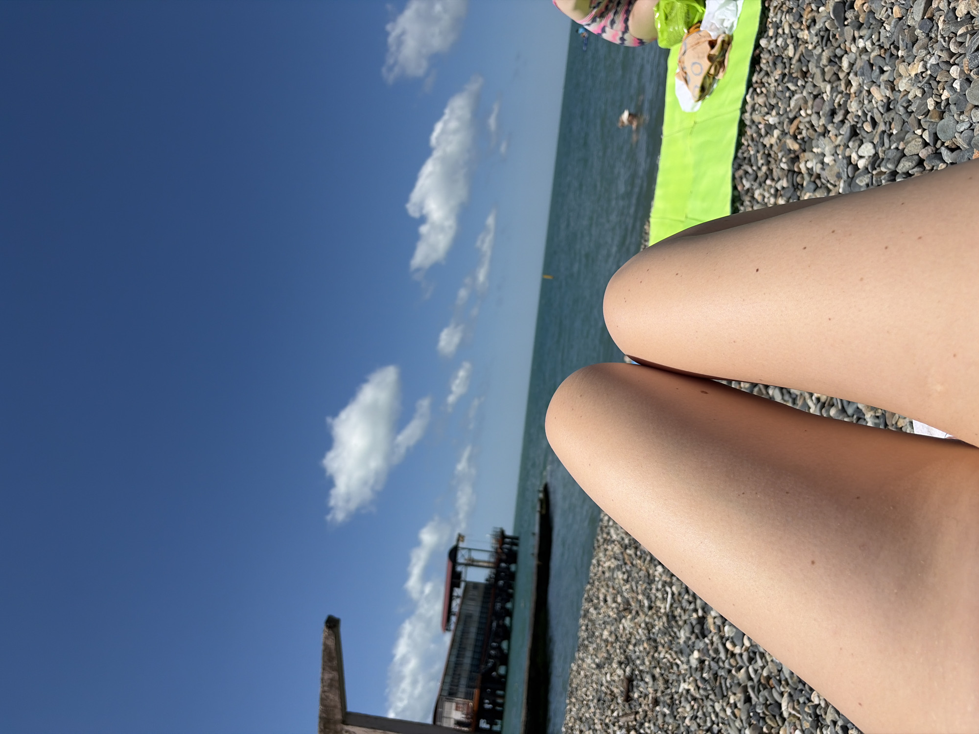 The image depicts a first-person perspective of someone relaxing on a pebble beach in Sochi, Russia. In the foreground, the subject's bare legs are prominently featured. The mid-ground shows a bright green towel laid out on the pebbles, with some food items and a white cloth on it. Further back, the Black Sea stretches out, with a few people visible swimming. On the left side of the image, an old pier or structure extends into the water, with a building at its end. The sky is a clear blue with scattered white clouds.