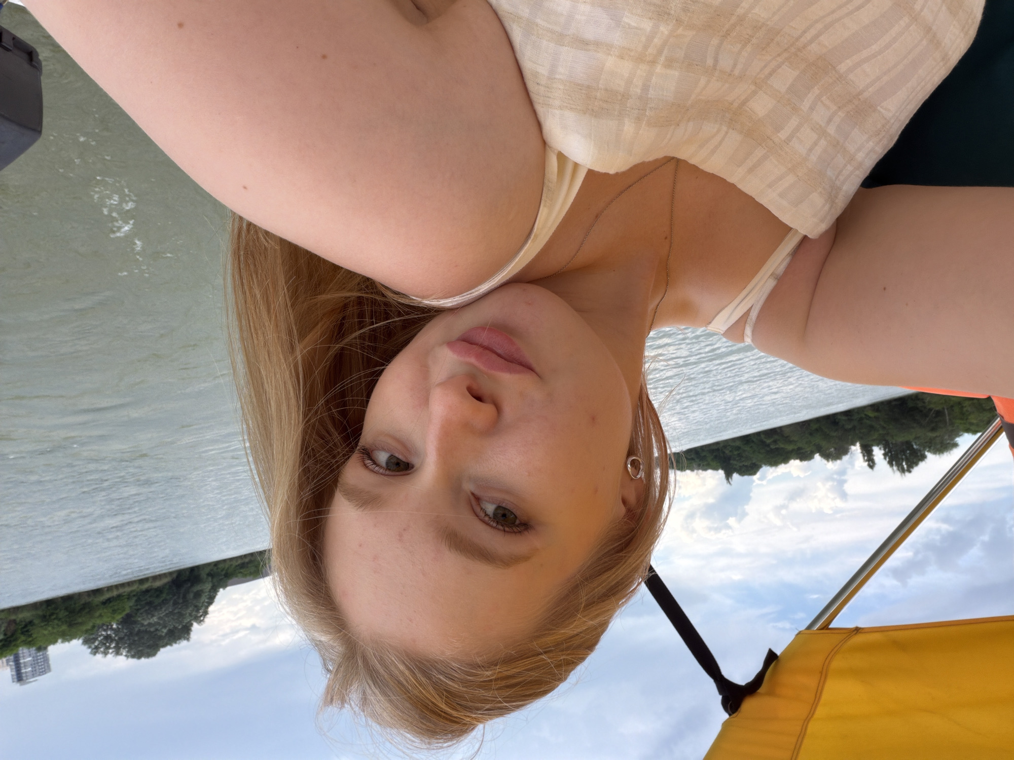 A young, blonde woman with fair skin is taking a selfie on a boat on a river. She is wearing a light-colored, plaid tank top and a delicate silver necklace. Her hair is long and blonde, falling over her shoulders. She has subtle makeup on, including light lipstick. She is wearing a small hoop earring in her left ear. The boat has a yellow canopy. In the background, there is a body of water, with a tree-lined shore and a tall building visible in the distance. The sky is partly cloudy. The setting appears to be a warm, daytime scene, likely in an outdoor recreational area. The location is the Kuban River in Krasnodar, Russia, given the presence of the specific building visible in the background, which is the "Onegin" residential complex located on the embankment of the Kuban River.