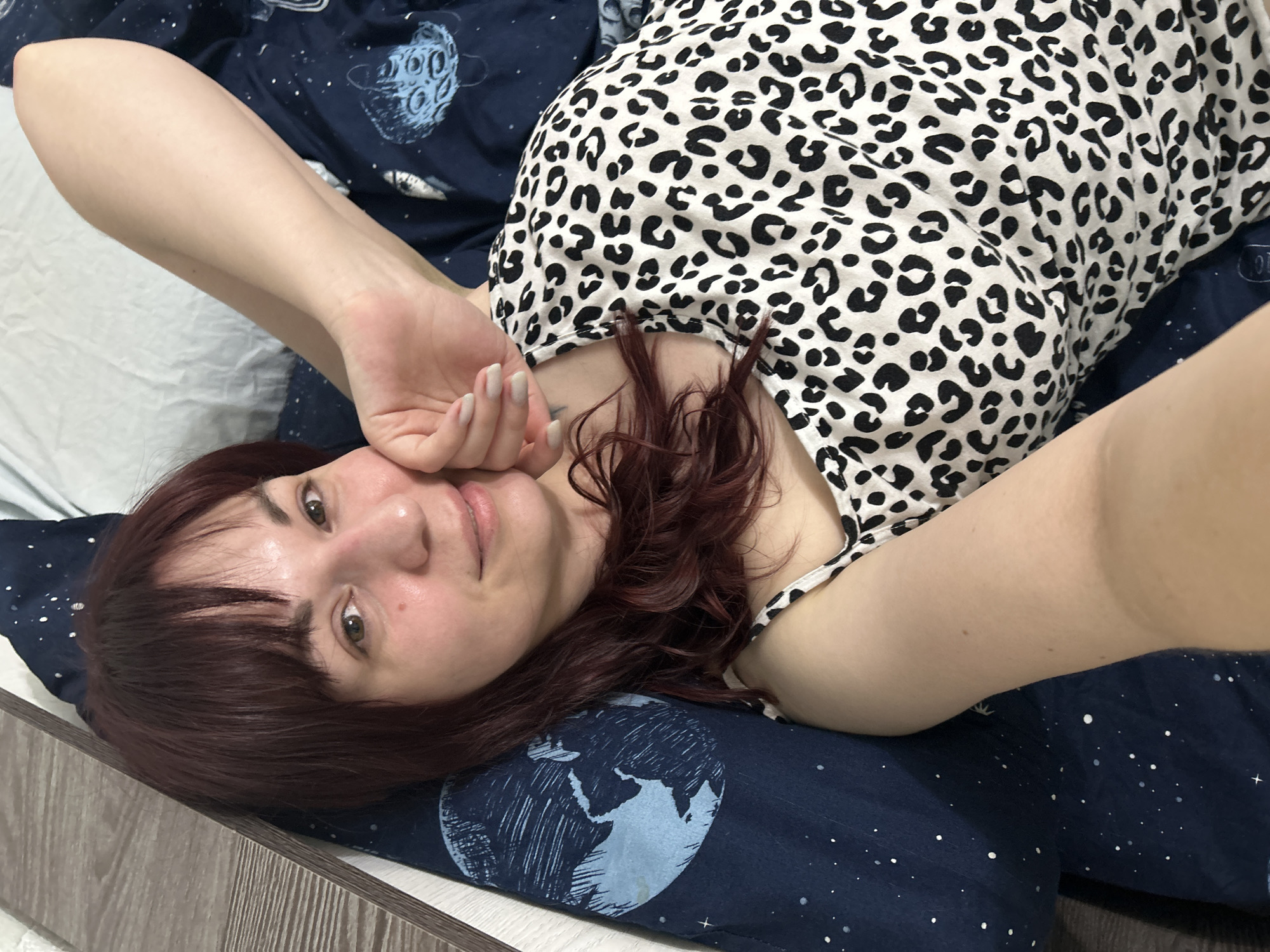 A woman with dark auburn hair and a white and black spotted top is lying on her back on a blue pillow with a globe and star pattern. She is holding her left hand near her face and smiling. The location is not discernible from the image.