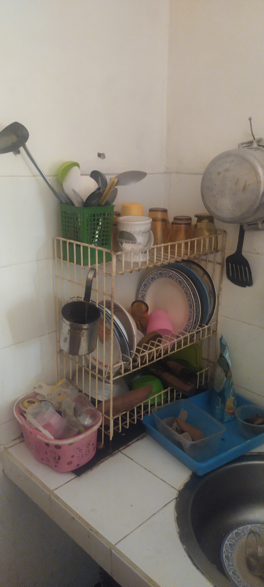 The image depicts a corner of a kitchen in Pasirhuni, Indonesia. The scene is cluttered with various kitchenware items organized on a three-tiered wire rack. The top shelf holds a green basket filled with utensils, including spatulas, spoons, and knives. Next to it are several amber-colored glasses and a white mug with text. The middle shelf displays a stack of plates, one with a blue and white pattern, and a pink plastic cup. Below this, a metal ladle hangs from the rack. The bottom shelf is occupied by a pink plastic basket containing baby bottles and other small items, along with a long, dark object that appears to be a vegetable. A blue plastic tray is placed on the countertop to the right, holding more kitchen items and a sponge. A large pot hangs on the wall to the upper right, and a black spatula is suspended below it. The countertop is tiled in white, and a sink is partially visible in the lower right corner. The overall impression is of a functional, albeit somewhat disorganized, kitchen space.